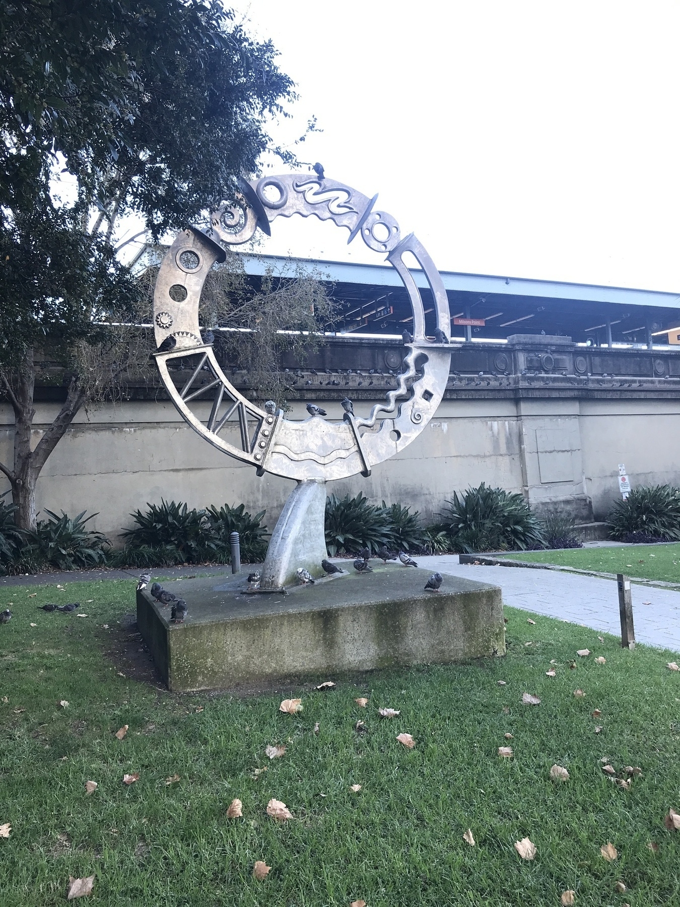 A circular metal sculpture in the shape of a ring, made from individual sections welded together, with a flat plate at each join. The sections have cutouts in swirly shapes, cogwheel shapes, spirals and ellipses, and one contains steel bars set in a zigzag pattern like a bridge. The main structure stands on a steel wedge shaped like a shark fin, which in turn is bolted to a concrete plinth. The sculpture stands on grass beside a path, with a wall and train station behind it. A pigeon sits on the very top of the ring. Three pigeons sit at the bottom, inside the ring, and others on two of the metal plates that lie horizontally. A dozen pigeons sit on the concrete plinth, and two on the grass. The sky is white - washed out.