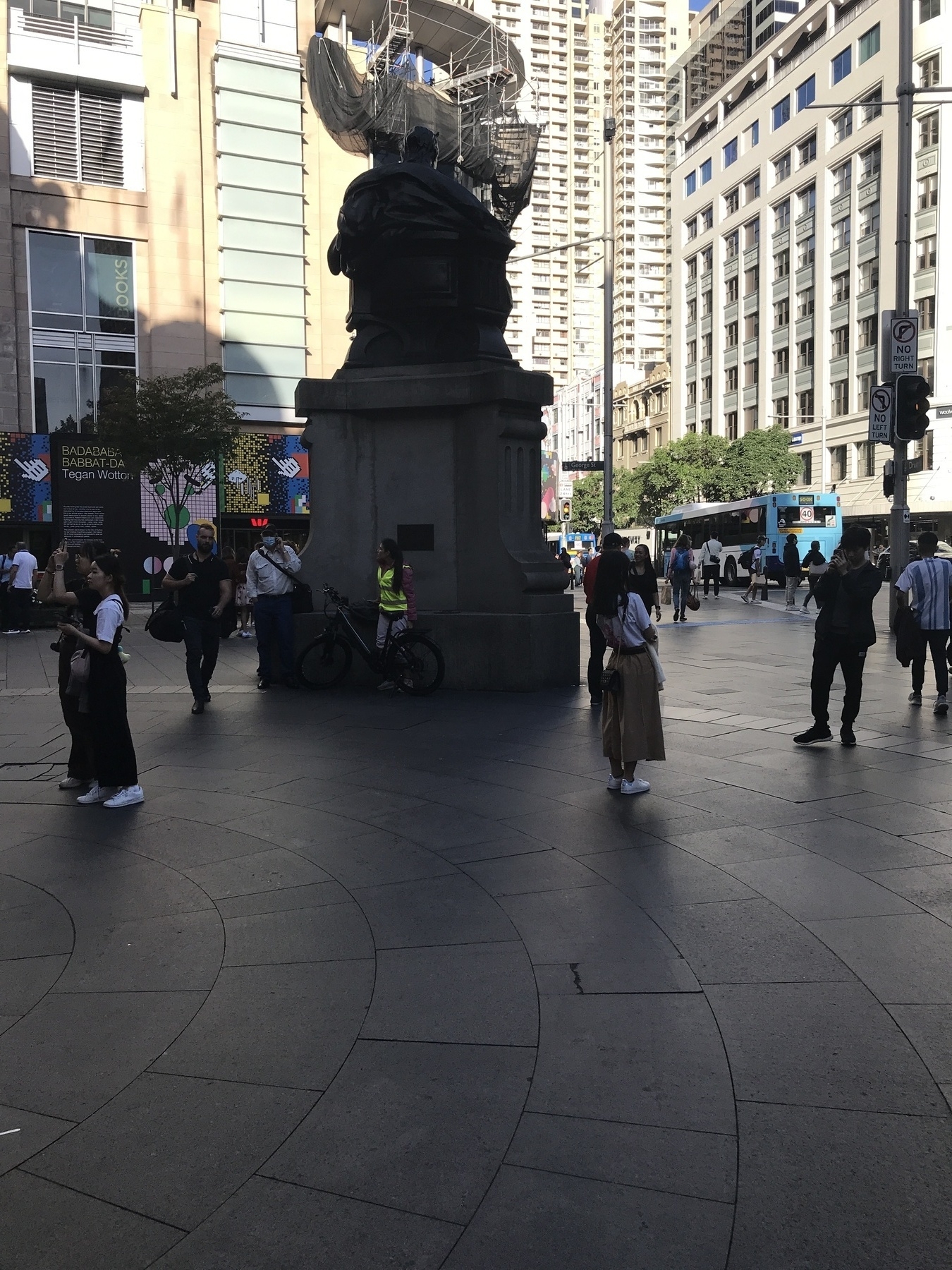 In the foreground is a shadowed open space with dark stone pavers laid in concentric circles. The space is dominated by a statue of Queen Victoria, seen from the back. The statue is in deep shade and stands in silhouette against a backdrop of tall buildings lit brightly by the late afternoon sun. The statue’s plinth is the height of two people, and a young woman in a bright yellow vest leans against it with her pushbike. She has long dark hair, long red sleeves and grey trousers, and is looking off to the side as if she’s seen someone she knows. A girl stands nearby being photographed, and various tourists, workers and window-shoppers populate the square and the street beyond. The statue on the plinth is a throne, with the back of the queen’s head visible above it. In the middle ground, a sunlit building supports a structure of scaffolding draped with huge loops of grey mesh like gathered curtains. It merges with the statue and almost seems to be part of it, or perhaps a ship that hangs in the air.
