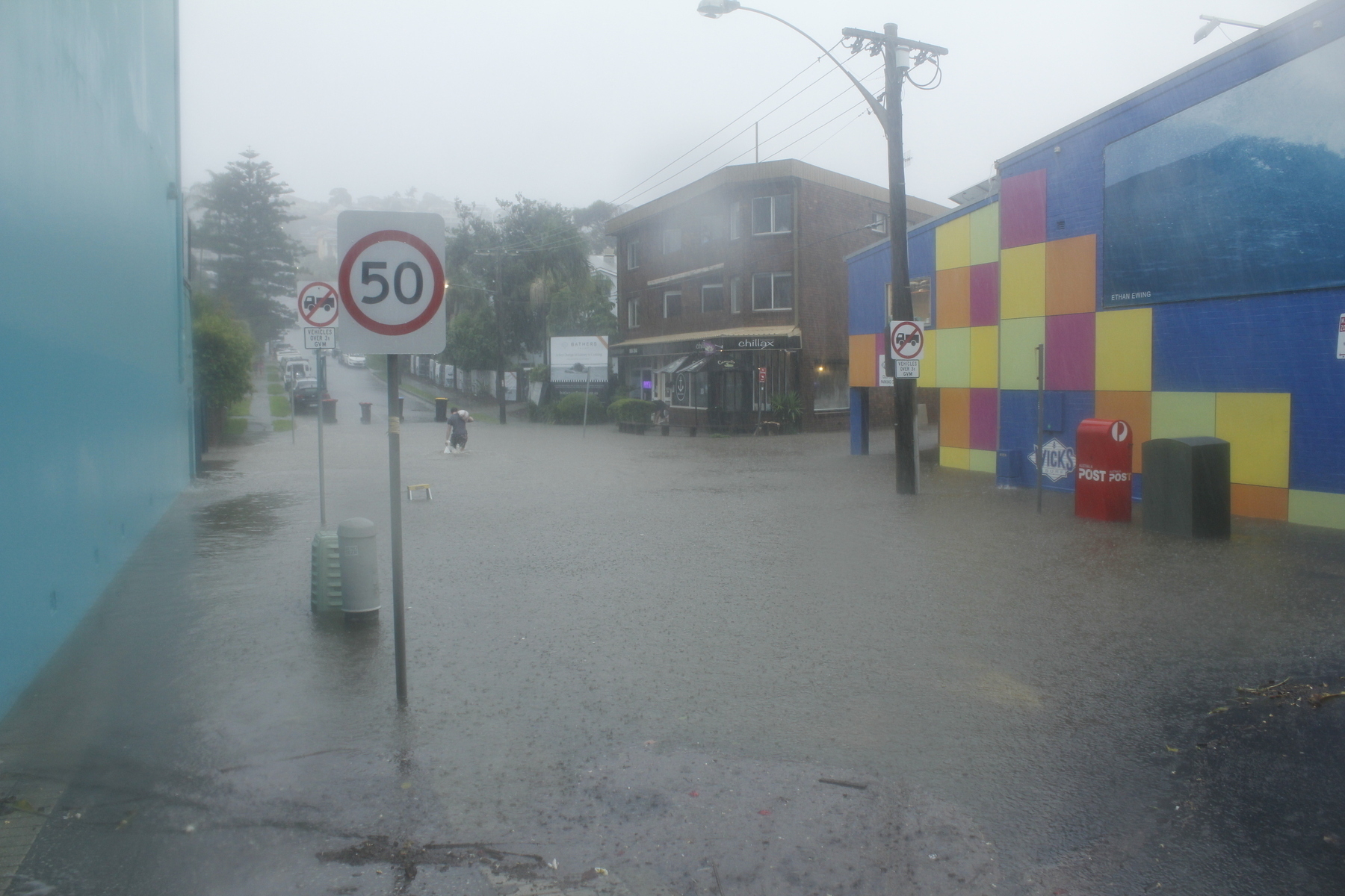 Looking into a street where grey floodwater covers the road surface and both footpaths, forming a lake in which stand traffic signs, street lights, telecom boxes, a street posting box and a step-ladder with only its very top showing above the water. Heavy rain falls on the lake, pitting its surface, and the sky and the ambient light are grey. In the distance a man slogs knee-deep towards a partly-submerged brick building. He has a sandbag on his shoulder, and clutches another bag that’s in the water. Outside the brick building a sign is displayed showing a woman lying on a beach, with the slogan: ‘Bathers - A Sea Change in Luxury is Coming’.