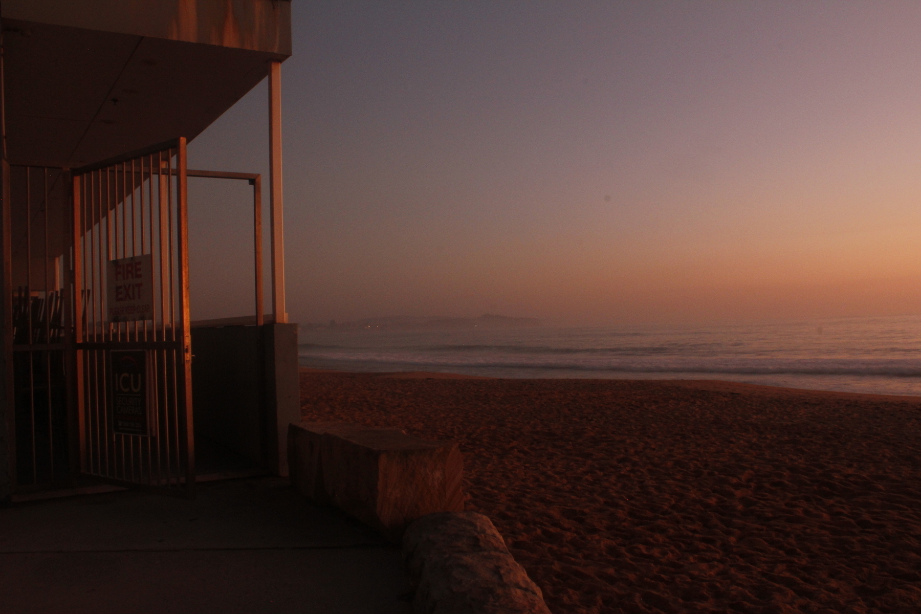 The edge of a sea wall and a building with an iron gate and open walkway, overlooking a beach and a small surf at dawn. The horizon is a fuzzy read with faint headlands fading into the distance. The beach and the building are lit with red and deeply shadowed. The sky is red, orange and purple.