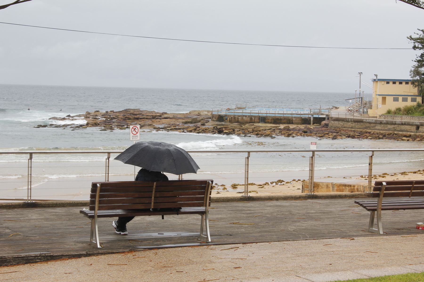 A lone figure, seen in silhouette from behind, sits beneath a black umbrella on a steel and timber bench overlooking the surf. The sea and sky are both grey, but the water is darker. Waves break on the tip of a rocky outcrop in the middle distance and sweep round onto the sand, which is littered with seaweed. A two-storey yellow building and a swimming pool extend out onto the outcrop. A silver railing separates the bench and walkway from a short drop down to the beach.