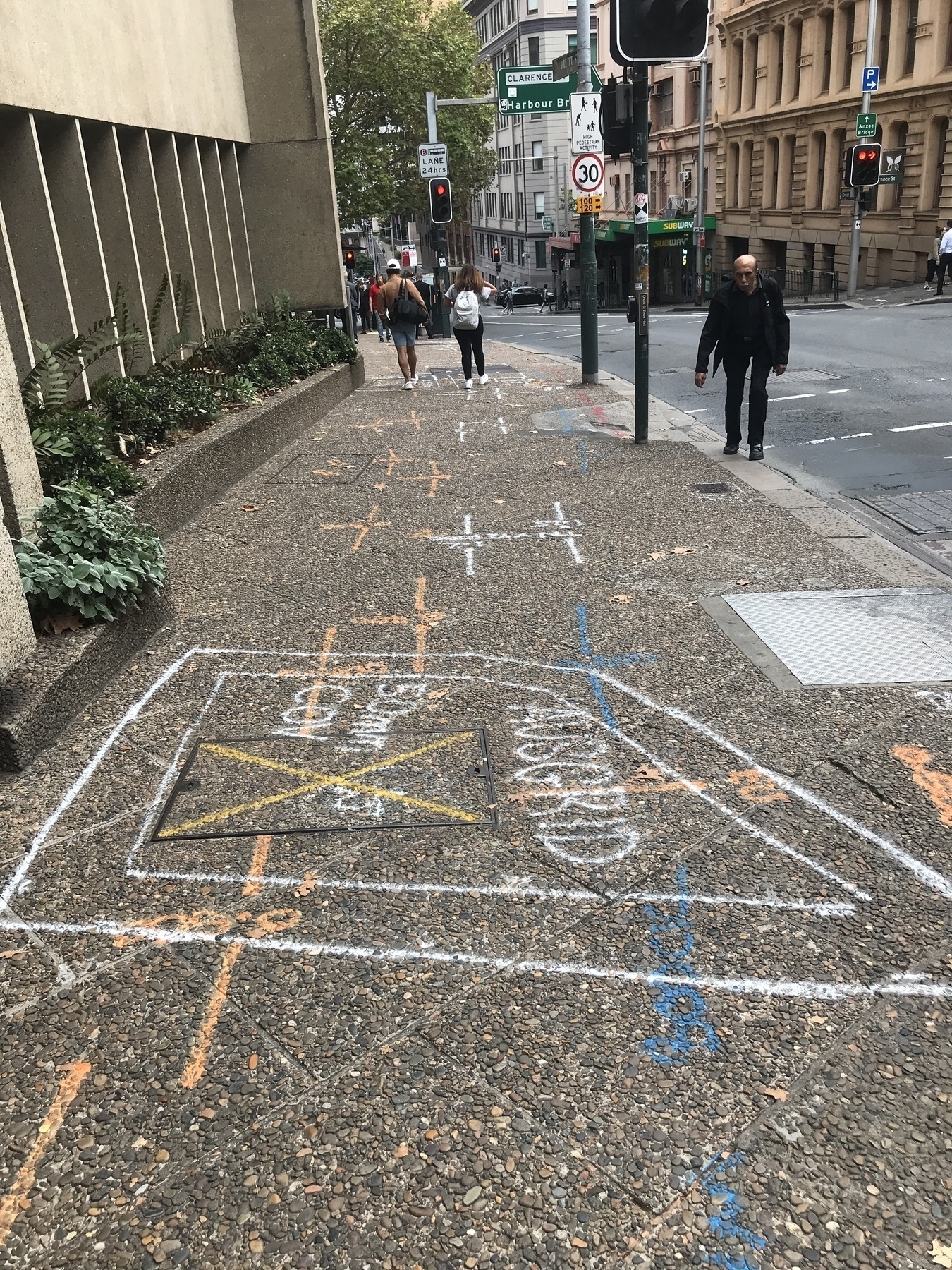 Beside a city street sloping sharply downhill, the wide footpath is covered with geometric shapes in orange, white, yellow, blue and red. Stick-aeroplane shapes in orange and white predominate, running down the slope in tandem, but there is are rectangles as well and lettering like 'Ausgrid' and '50mm Cov'. A stooped man in black looks uphill at the camera.