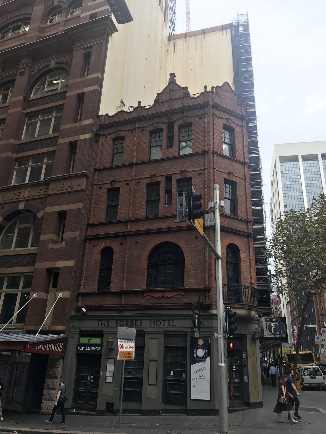Looking across King Street Sydney at the side of the Forbes Hotel, a four-storey brick structure with ornamental brickwork on the top three floors. An undulating half wall at the top, incorporating two tall triangular peaks, is very clear against two cream walls of a taller building behind. Every door and window is black timber and glass. The ground floor is half grey wall and half black doorways, with the hotel name at the top.The hotel presses against a taller building next door which is also brick, and shows the lettering 'Warehouse 1914'.