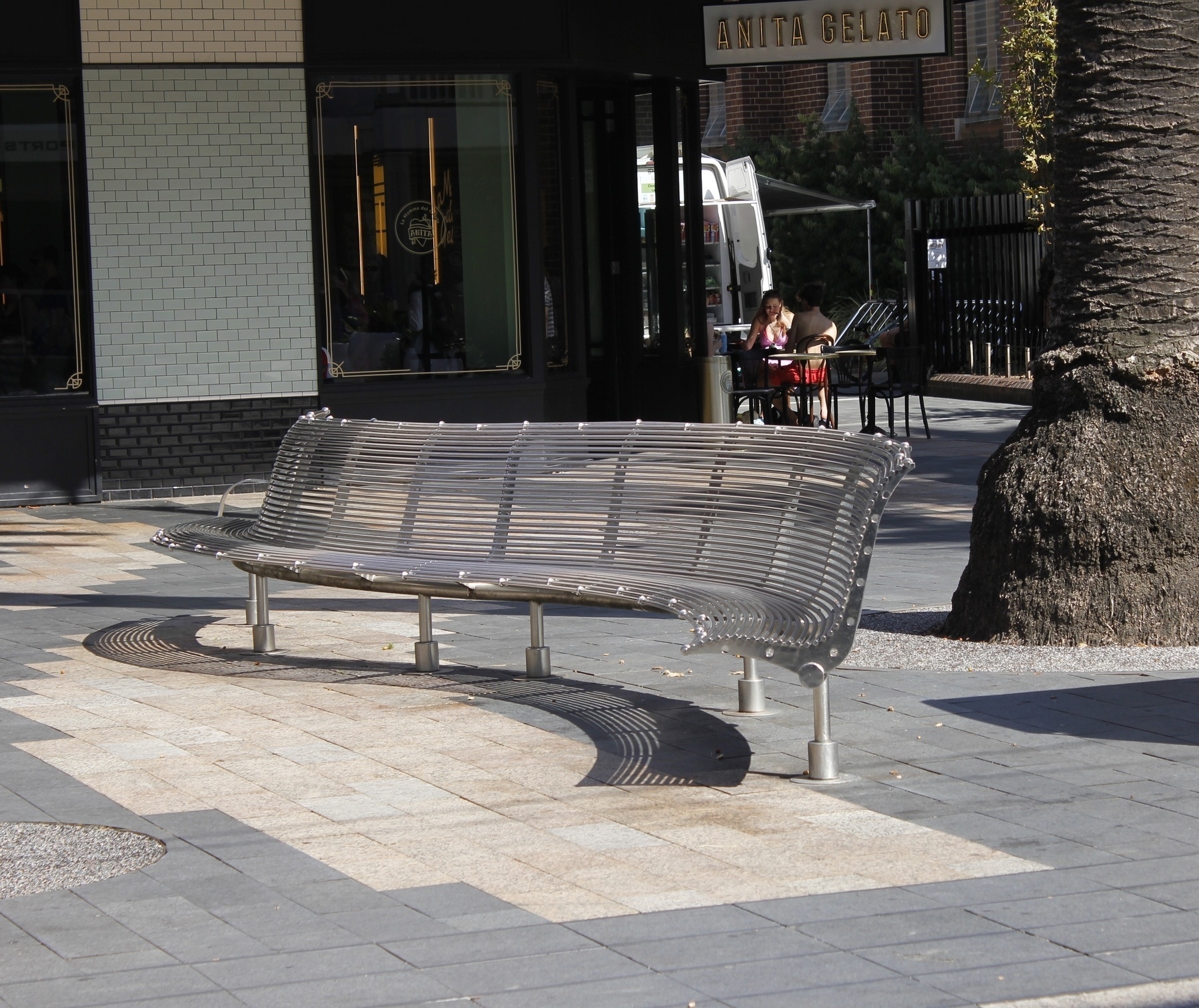 A steel-coloured metal bench stands on six steel legs, on a sunlit, tiled footpath in the foreground. The bench curves at the ends, in opposite directions, so that its shadow on the footpath forms a swirling, elongated letter 'S', in shadow lines due to the many narrow, horizontal plates that form the back and base of the bench. A palm tree stands behind the bench. In the background, in deep shadow, is a shopfront with a sign saying 'Anita Gelato', and two people at a table outside.