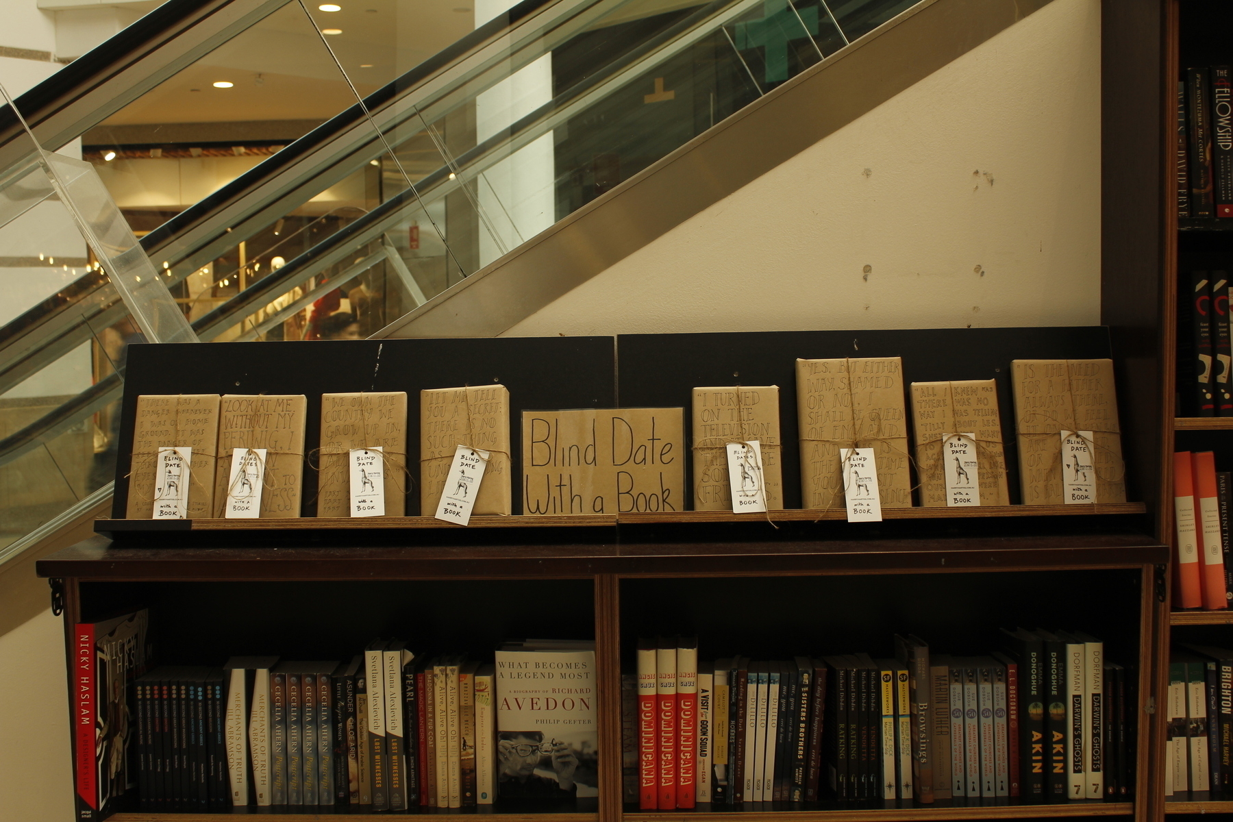 Two shelves of a book display against an escalator. A sign in the middle of the top shelf says ‘Blind Date with a Book’. On either side, facing out, are books wrapped in brown paper and tied with string, with a passage from each book written on the outside.