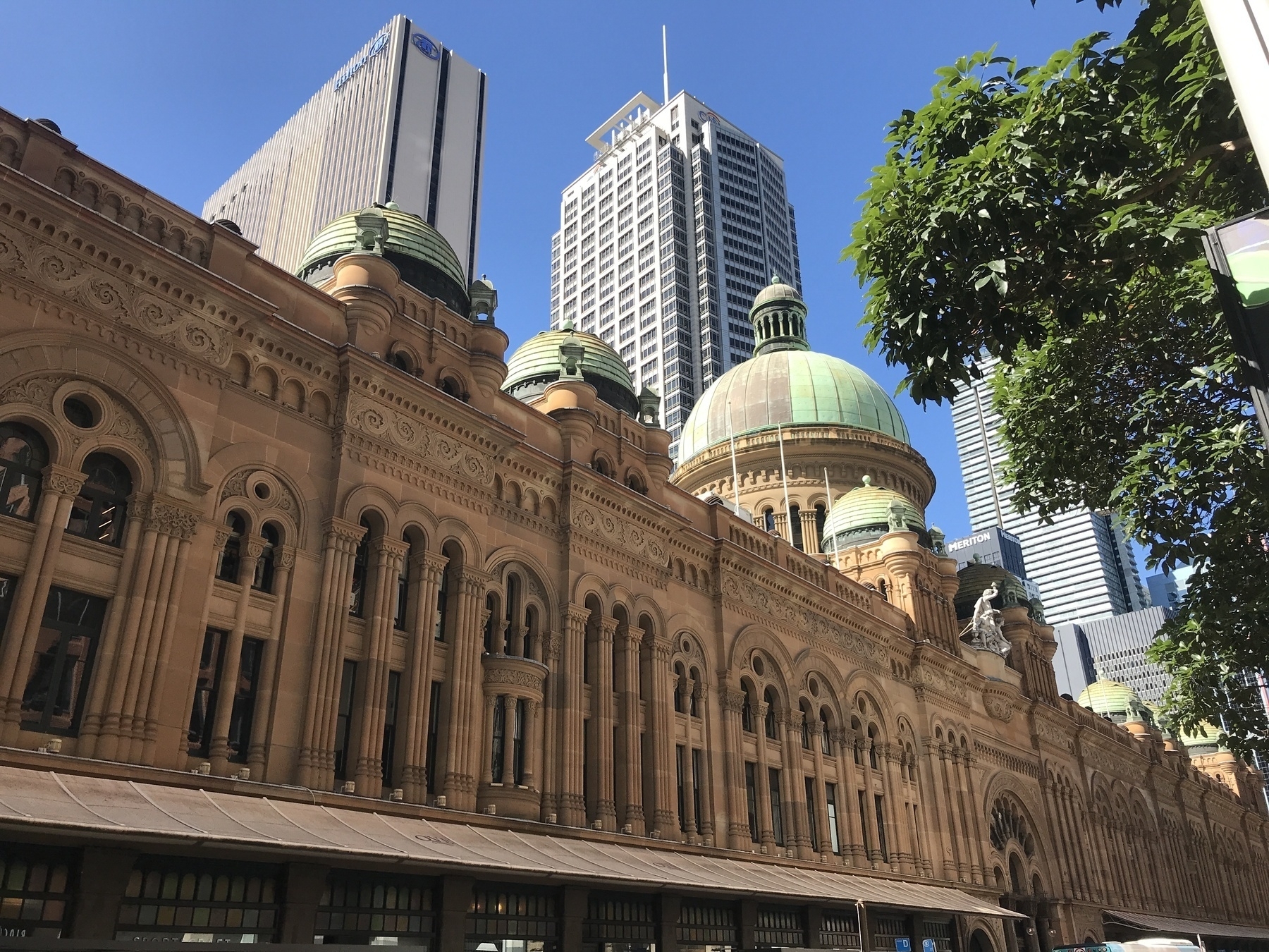 A long building made of sandstone stretches into the distance, topped with copper domes that have aged to green. The ground floor is all windows beneath a light and graceful awning. Above this are many ornate arched facades rising two storeys, bisected by horizontal stone bands that separate the lower, tall, rectangular windows from the shorter arched windows above. The wall continues above these with bands of high and low relief, rising to a separate tier supporting the towers topped by copper domes. Tall, modern buildings rise in the background against a bright blue sky.