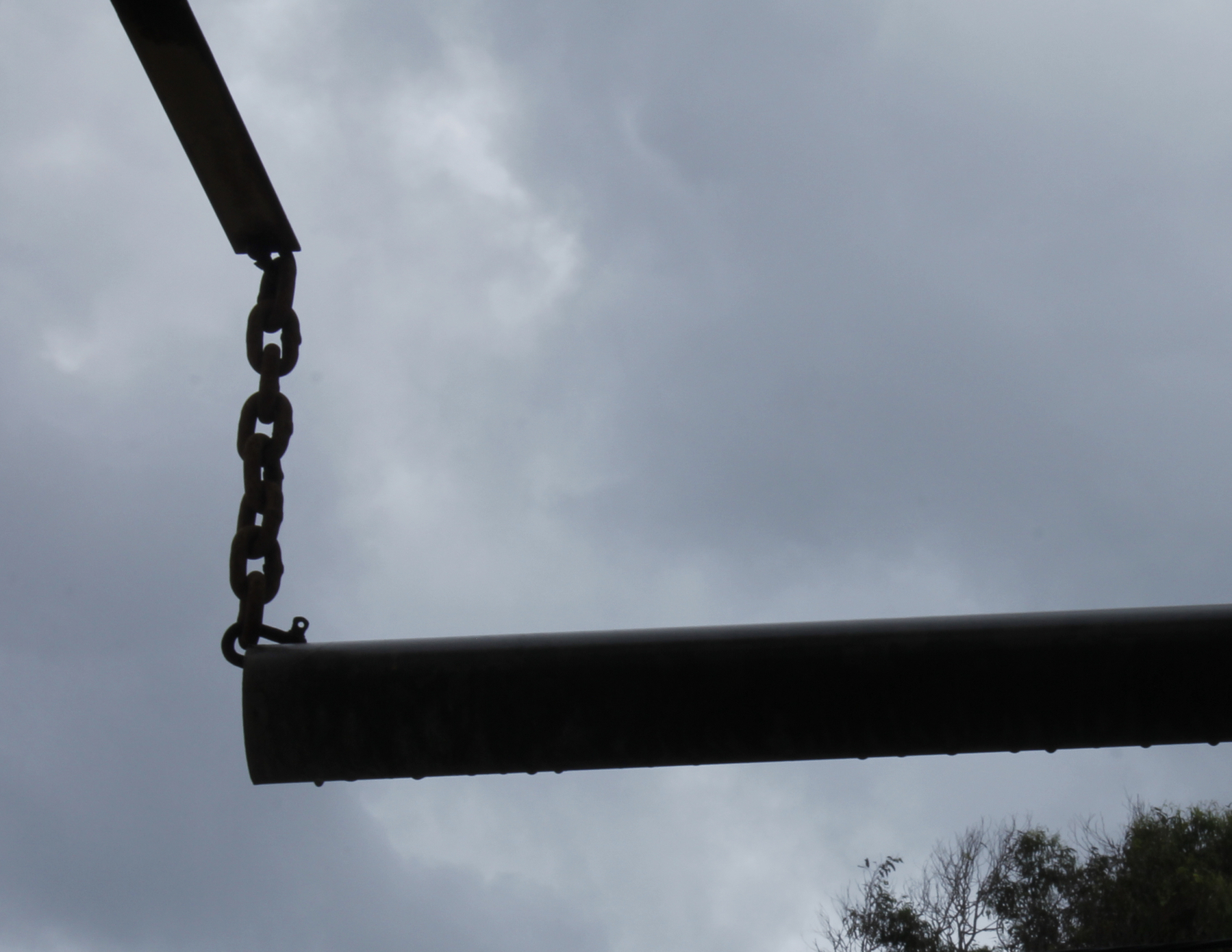 A horizontal pipe is silhouetted against a sky of dark grey storm clouds. The pipe hangs from a short chain, which in turn is suspended from a metal bracket near the top left corner. The chain and bracket are also in silhouette. The bracket, chain and pipe are part of a low-tech mechanism to warn drivers if their vehicle is too high to enter the space beyond.