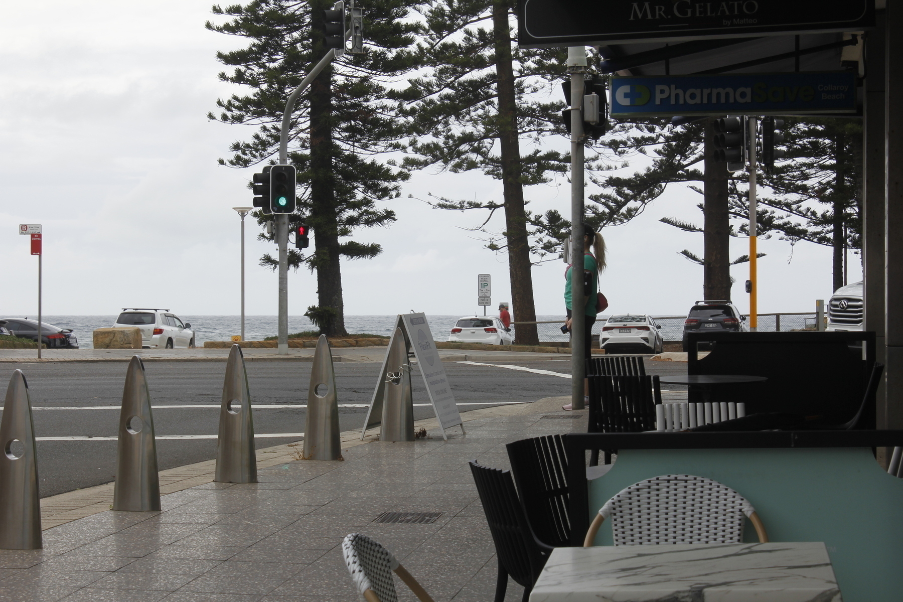 A street corner with bicycle stands and cafe chairs and tables in the foreground, palm trees across the road and the ocean beyond. The sky is a mass of pale grey cloud and the sea is dark grey and choppy. In the middle ground, on this side of the road, a woman stands facing left behind a pole, waiting to cross the side street; her ponytail spikes out behind the pole and her nose pokes out in front of it. The front and back of her body are on either side of the pole. In the distance, across the road, the timber top of a fence runs along the horizon from the right and dips below it in the middle of the image.