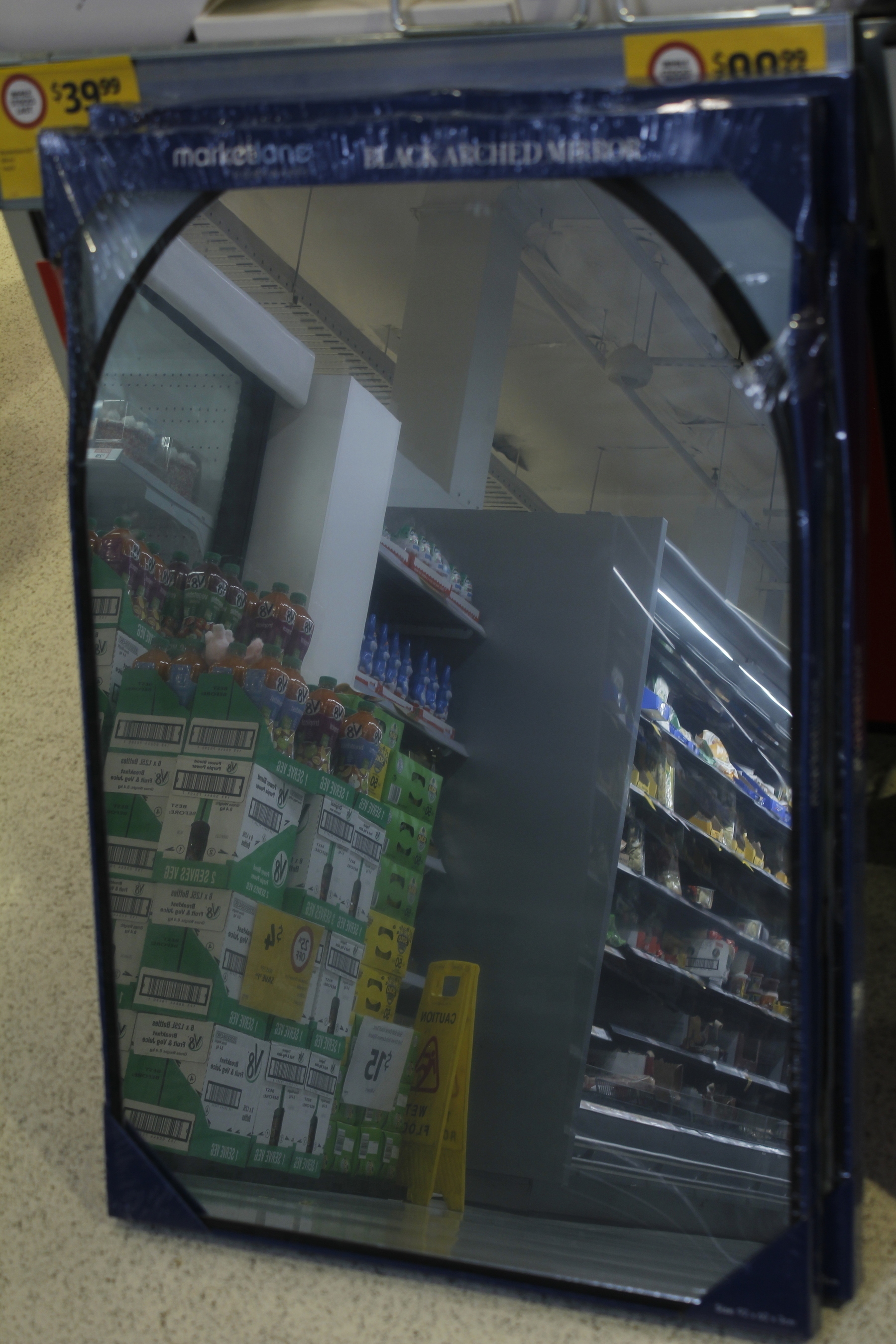 An arched, black-framed mirror stands in its packaging on a speckled floor, leaning so that everything reflected is also on an angle. Inside the frame, in reverse, are green boxes of V8 drinks stacked up at the side of a fridge, two shelves containing Easter eggs and chocolate Easter bunnies, and a big grey rack of open, refrigerated shelving full of boxes and packets lit by fluorescent tubes. A wide, straight-sided metal flue rises to a grey industrial ceiling with sprinklers, hanging lights and other attachments.