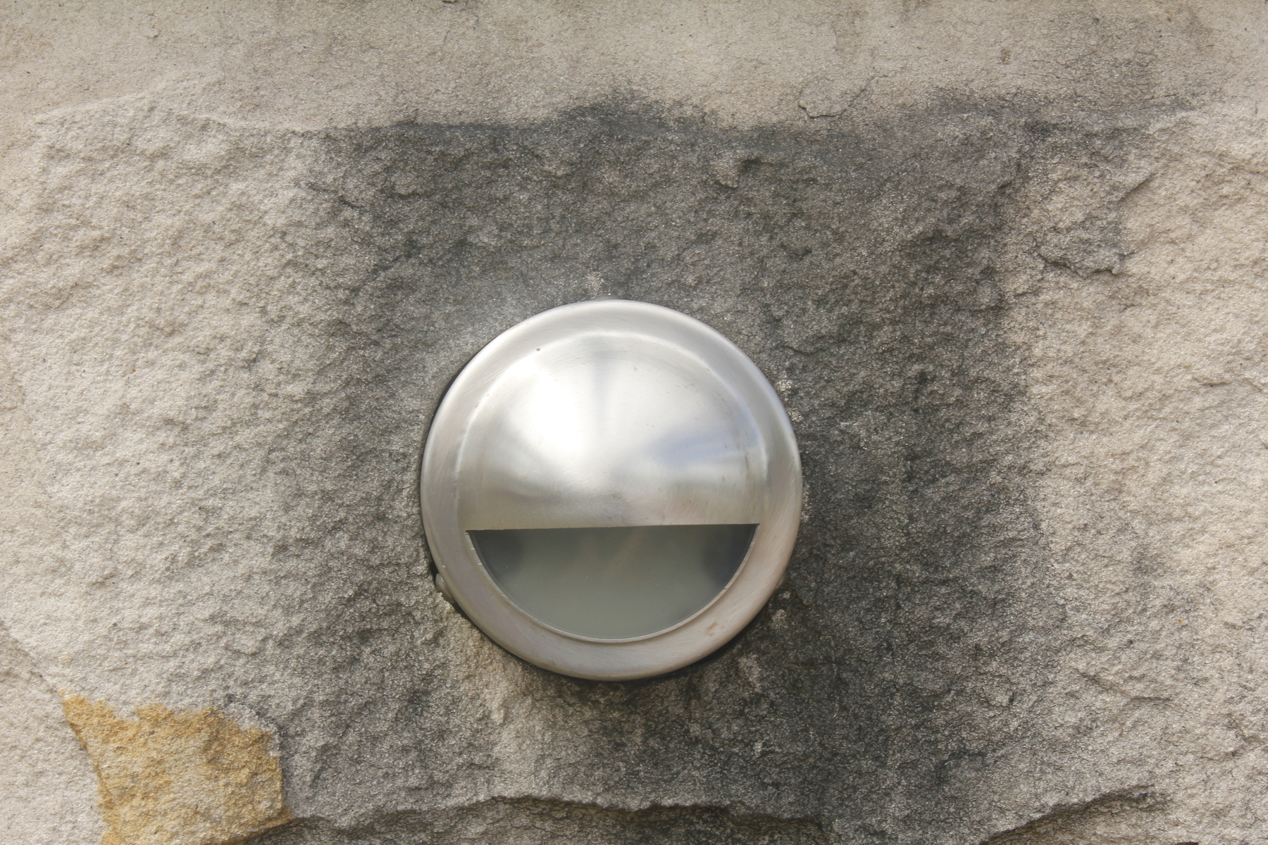 A shiny silver light fitting set into a stained sandstone block. The fitting is circular, with the top part curving outwards, and the bottom section cut out so that it resembles a mouth stretched wide in a big, happy grin.