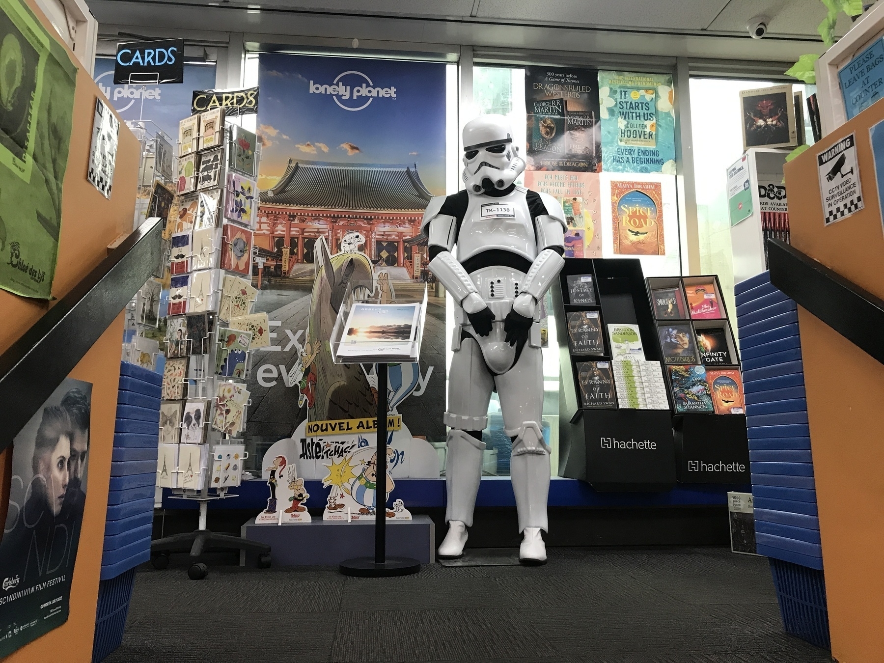A life-size Star Wars startrooper stands at the top of a staircase, surrounded by book displays, a greeting card stand and posters. Beside the trooper is a pile of catalogues in an angled tray. The posters feature ‘Lonely Planet’ guides and current fiction titles like ‘It Starts with Us’ and ‘Spice Road’, and an Asterix title lebelled ‘Nouvel Album’ in French, with cutouts of characters below.