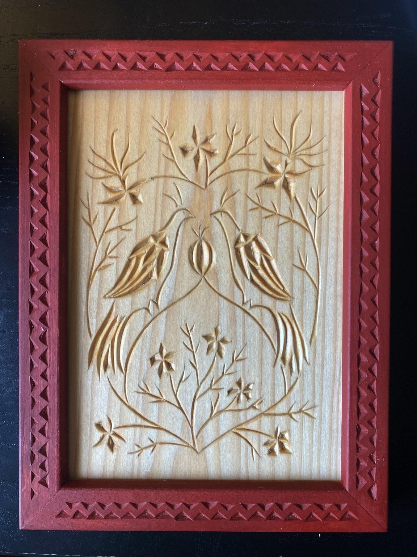 chip carving of two birds facing each other over unopened blossom; vines encircling them in the shape of a heart; many flowers; red-painted frame carved with woven triangles