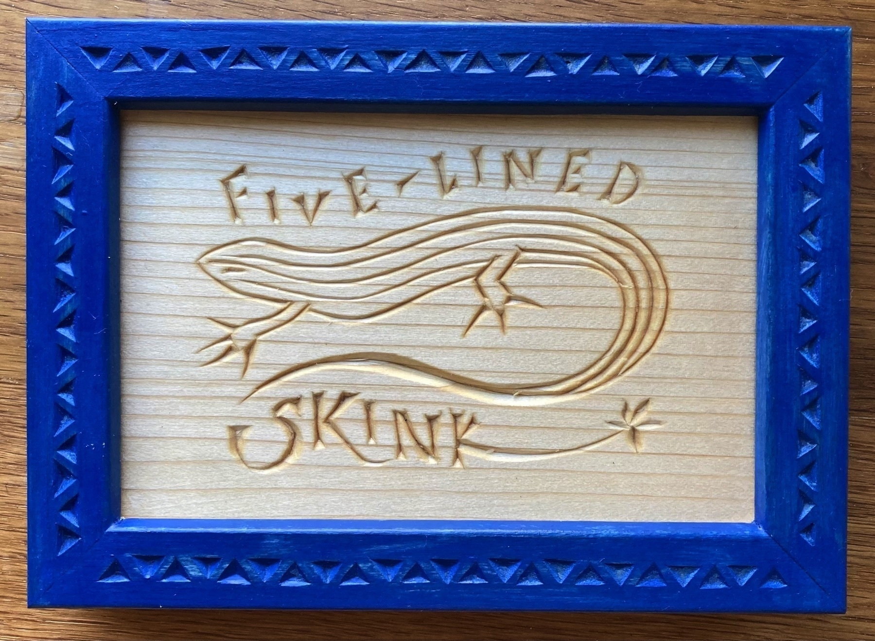 chip carving of a five-lined skink with caption, blue frame carved with triangles