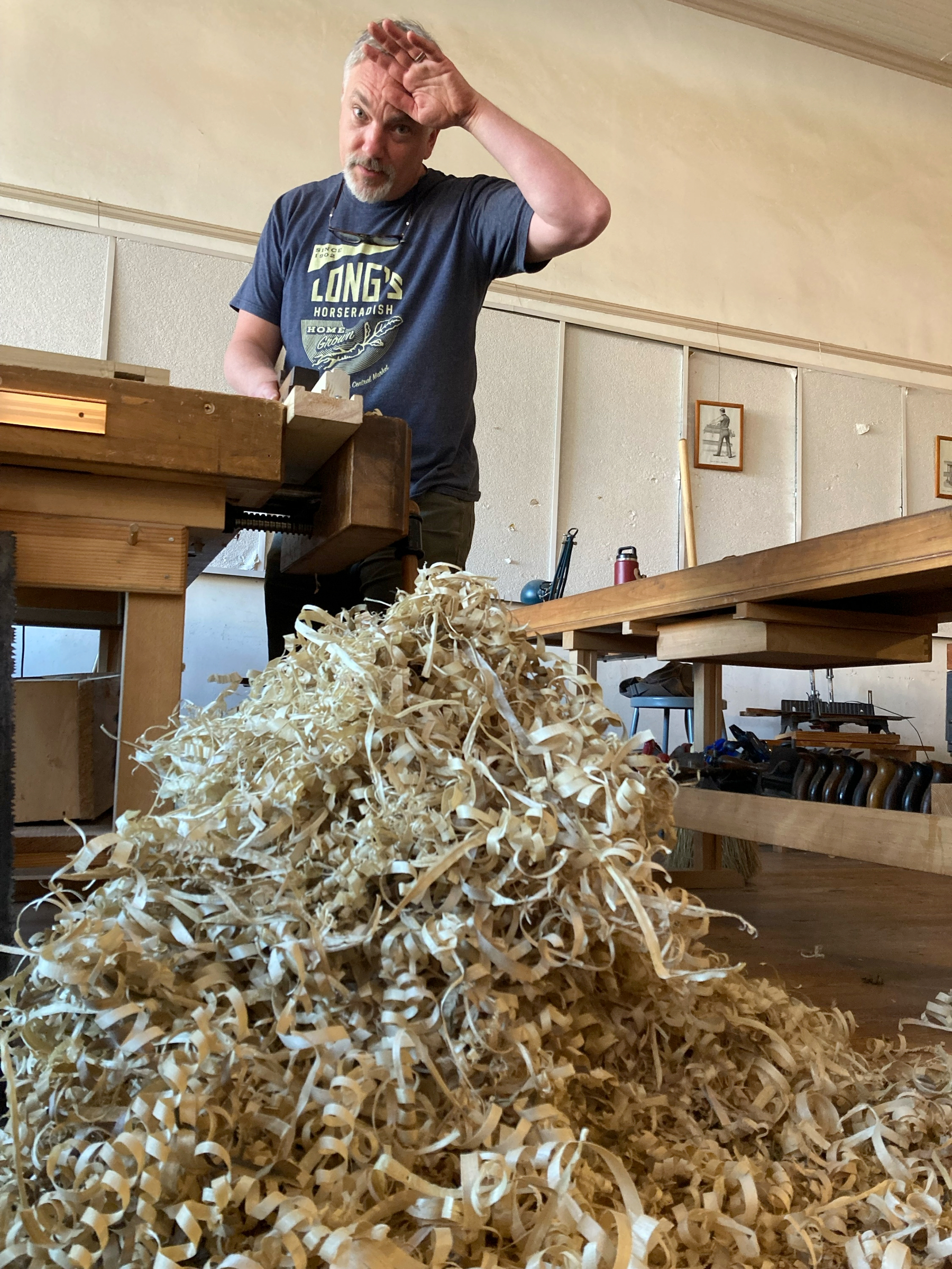me, faking visible exhaustion, standing at a workbench over a massive pile of wood shavings