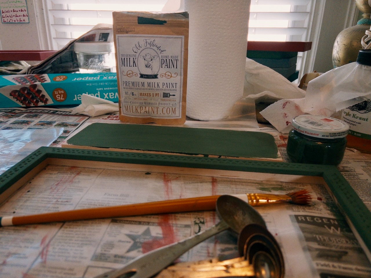 packet of powdered milk paint, small jar, paintbrush, measuring spoons, and a green-painted frame