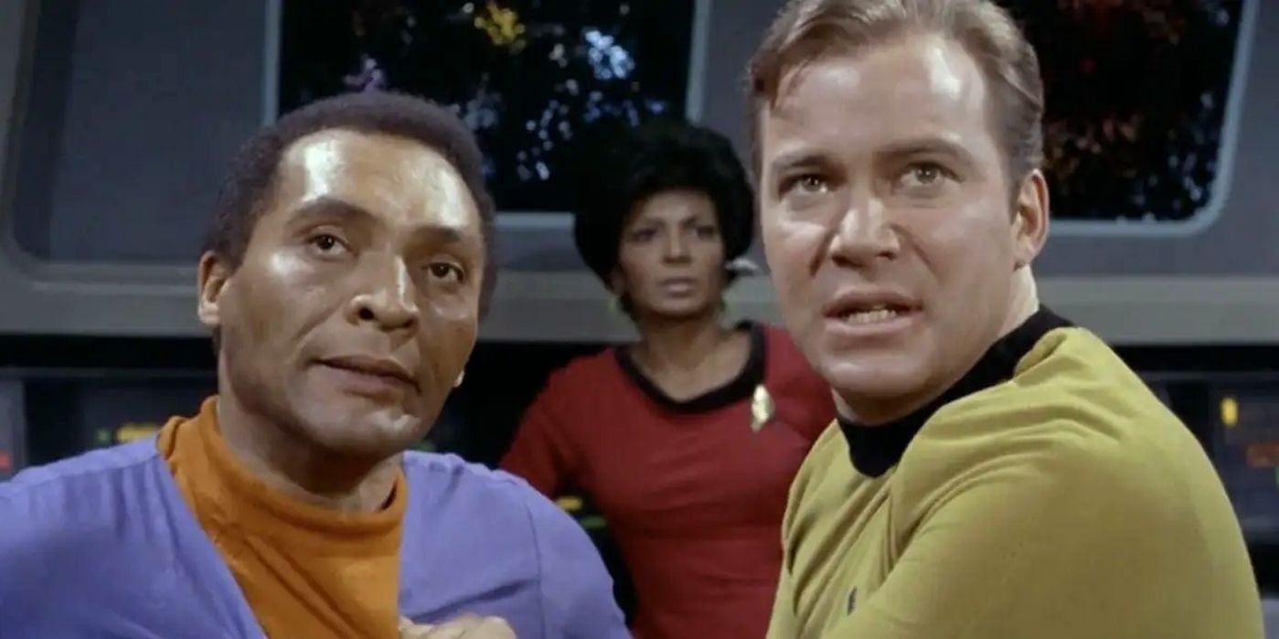 Two individuals in blue and yellow uniforms are in a tense situation on a spaceship's bridge, with a woman in a red uniform in the background. Captain Kirk is on the right, Uhura in the middle, and Dr. Richard Daystrom on the left.