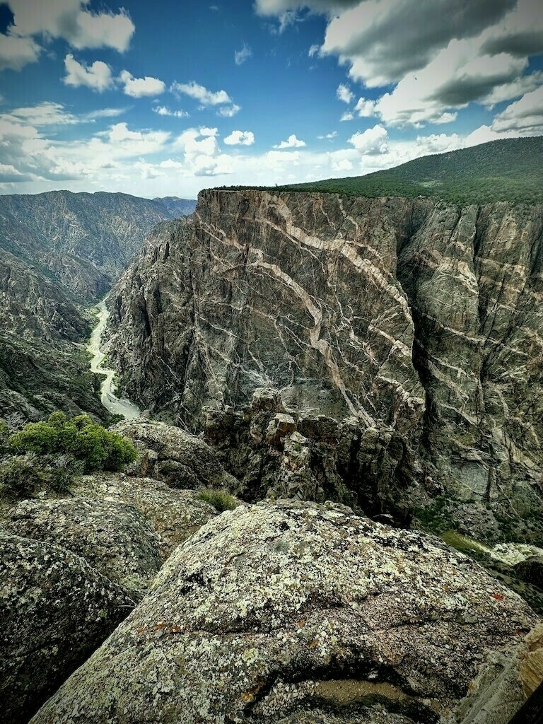 View of Black Canyon of the Gunnison's painted wall. Blue sky. Clouds. Winding river below. 