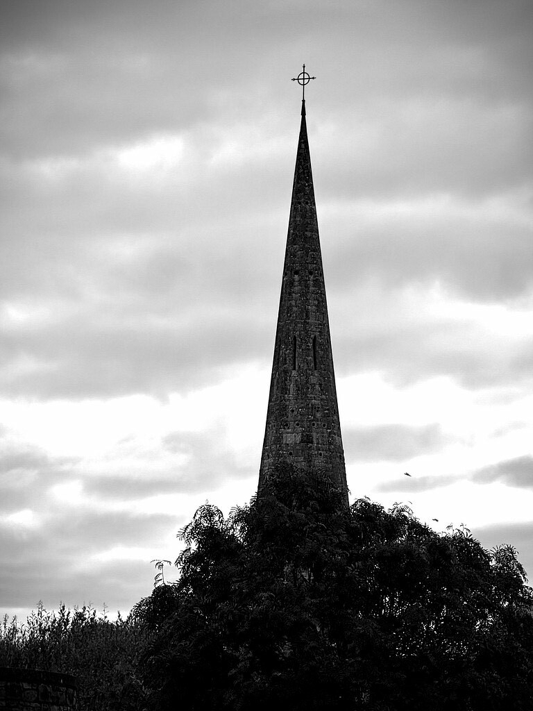 Very tall and narrow church steeple with cross on top against a cloudy sky. Black and White. 