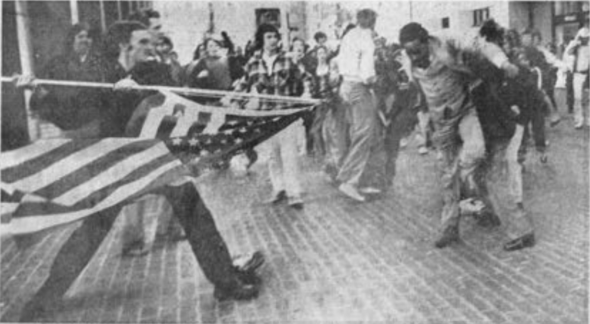A black and white photograph of a Black man in a plaza being attacked by a White teenager brandishing the American flag