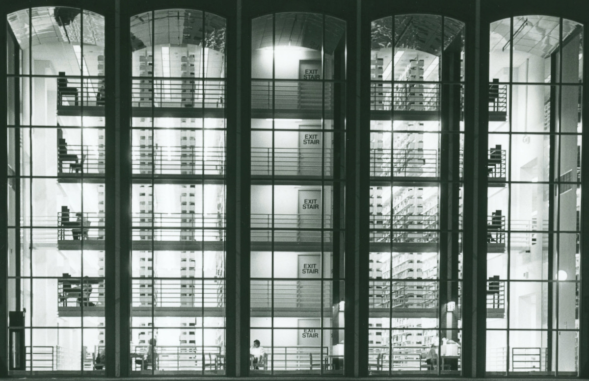 A black and white photograph of a library at night from the outside, with large glass windows showing the stacks and people studying