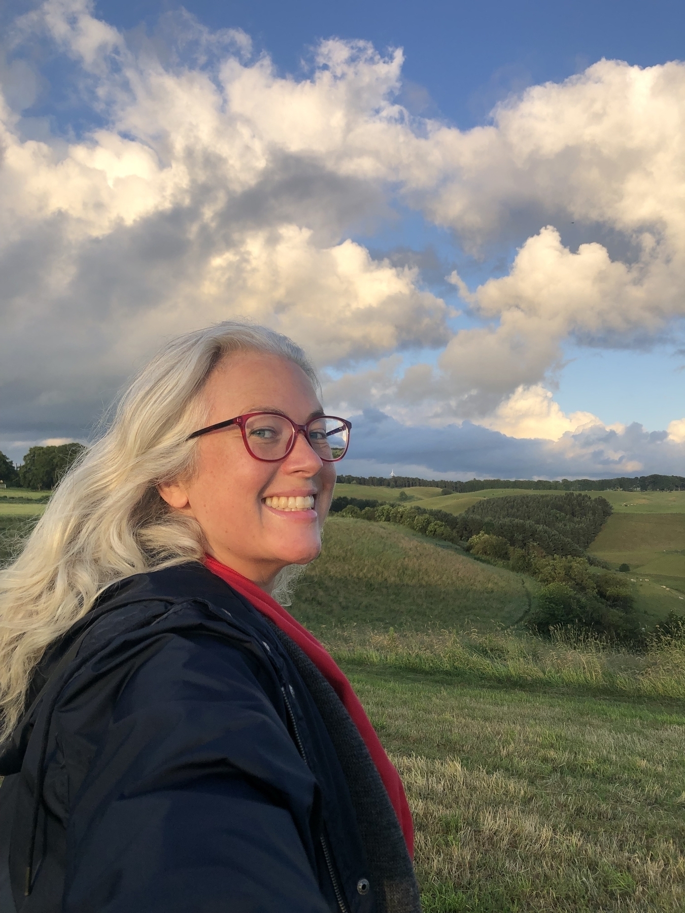 Sunlit clouds, green hills and a girl with red glasses and silver hair smiling profusely