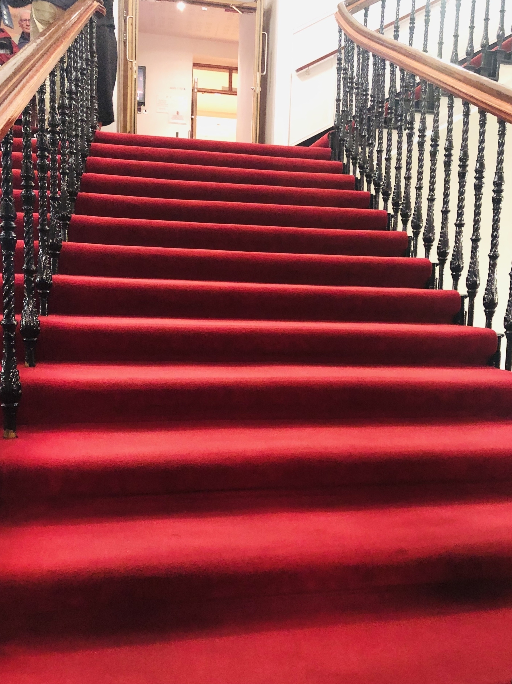 Ascending, grand red carpet of stairs with a wooden balustrade and black iron railings. 