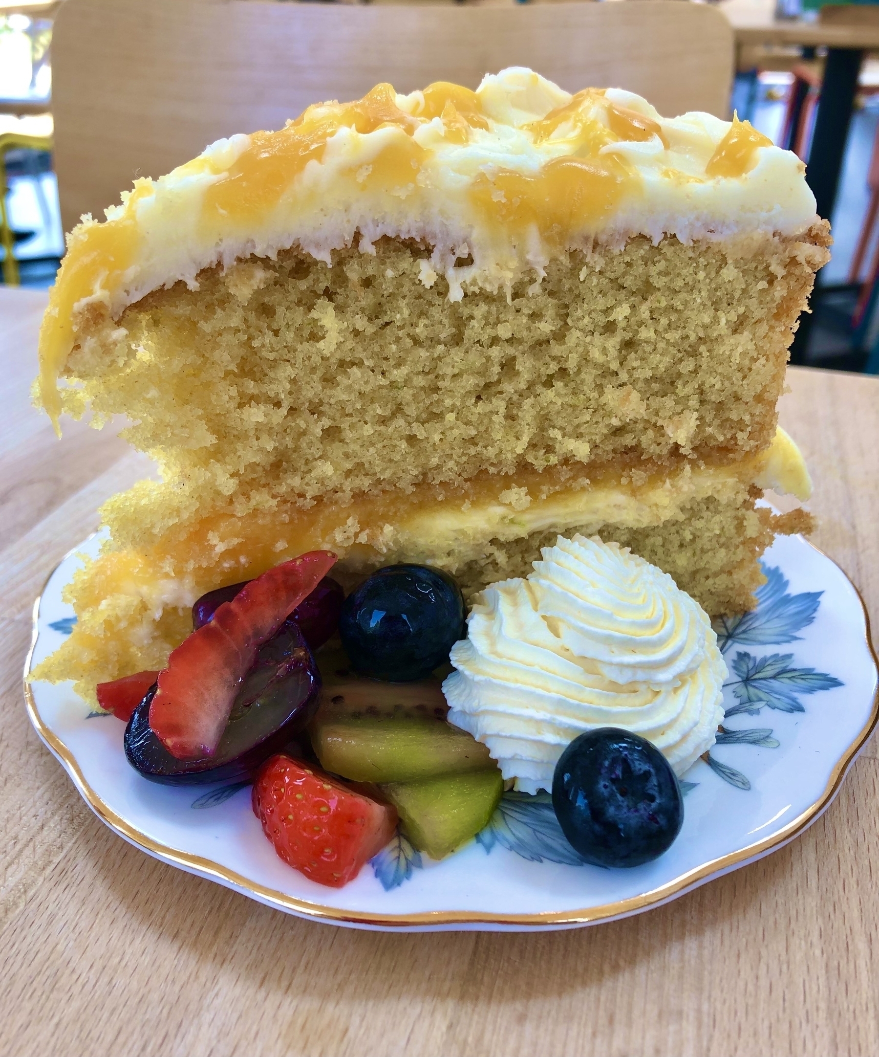 A slice of lemon sponge cake on a delicate bone china plate accompanied by fruit and cream. 