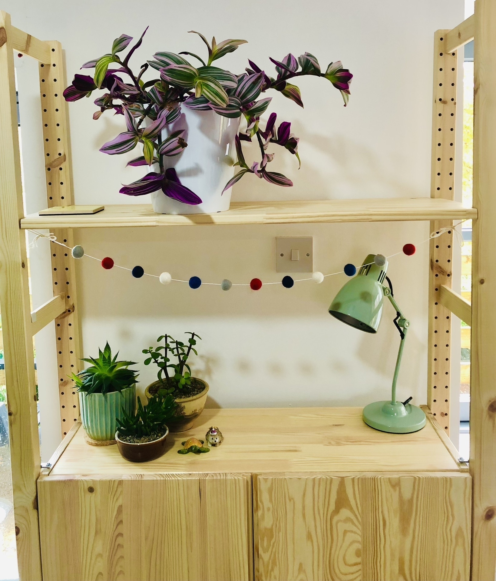 a pine cabinet with various plants on top and a string of colourful pom pom bunting underneath the shelf.
