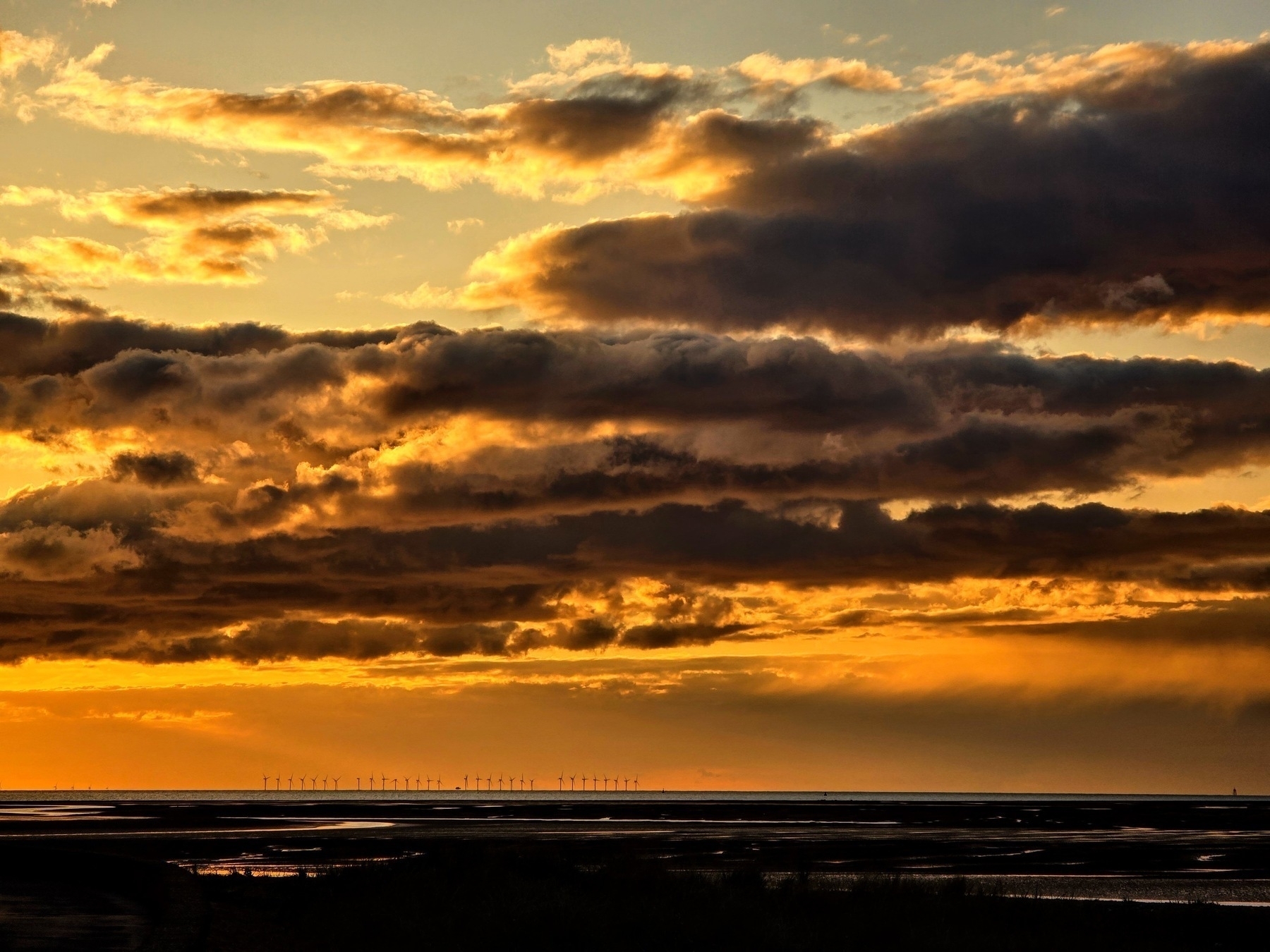 sunset os orange and yellow over a bay, the tide is out, wind turbines stand on the horizon