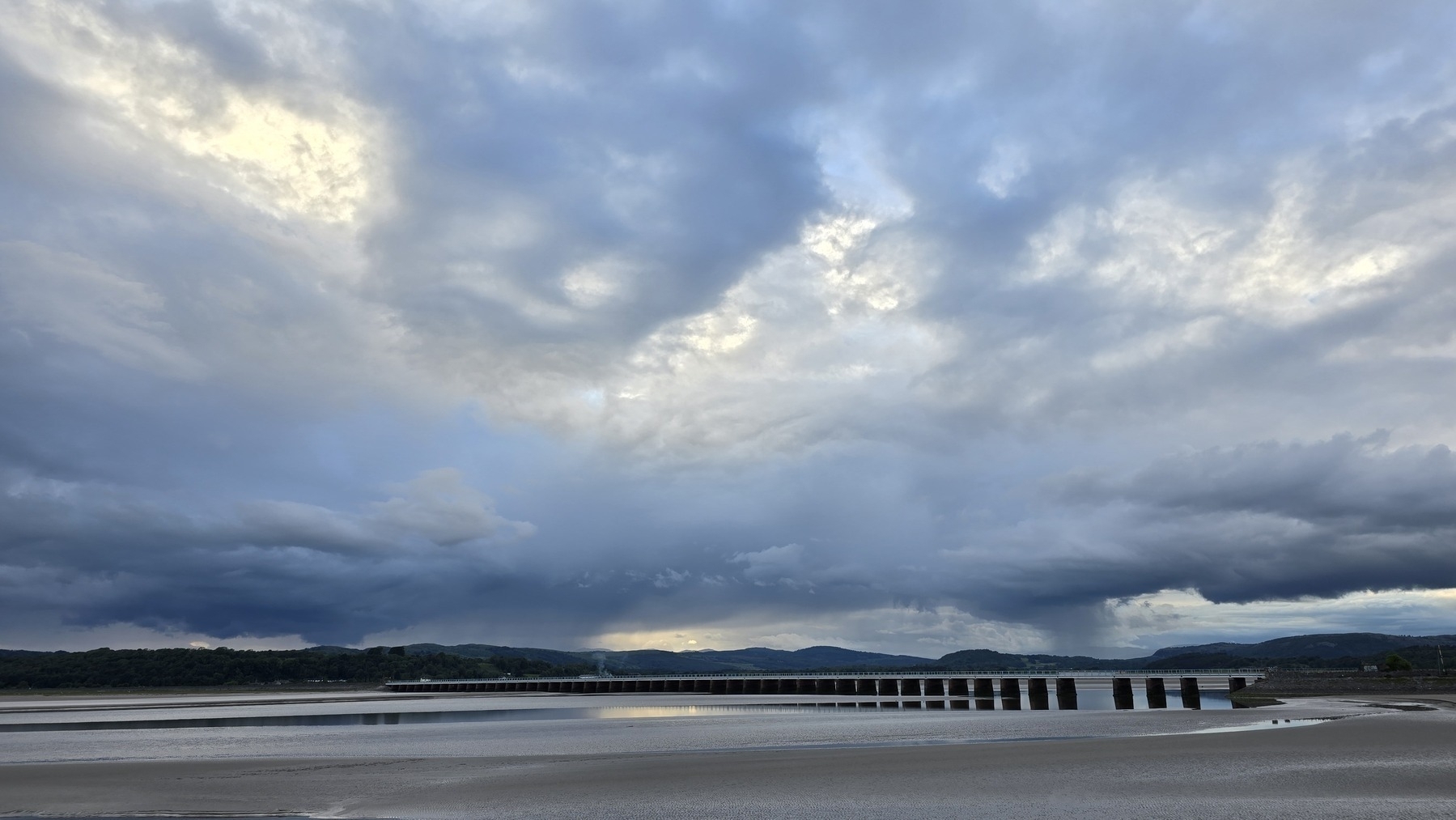 Landscape of a swirling, blue and grey cloudy sky taken over an estuary with a train bridge in the distance