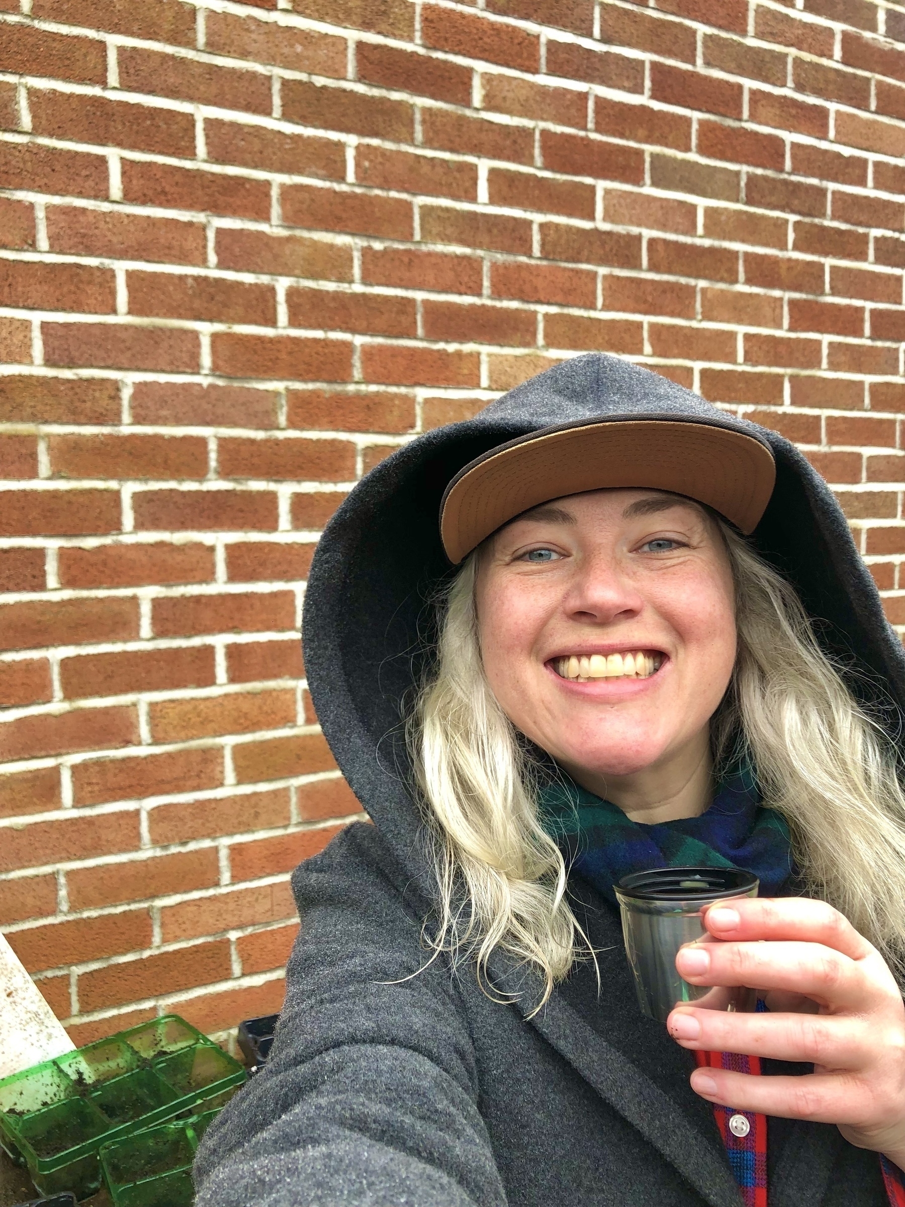 Holly drinking tea in a black hoodie in front of a red brick wall