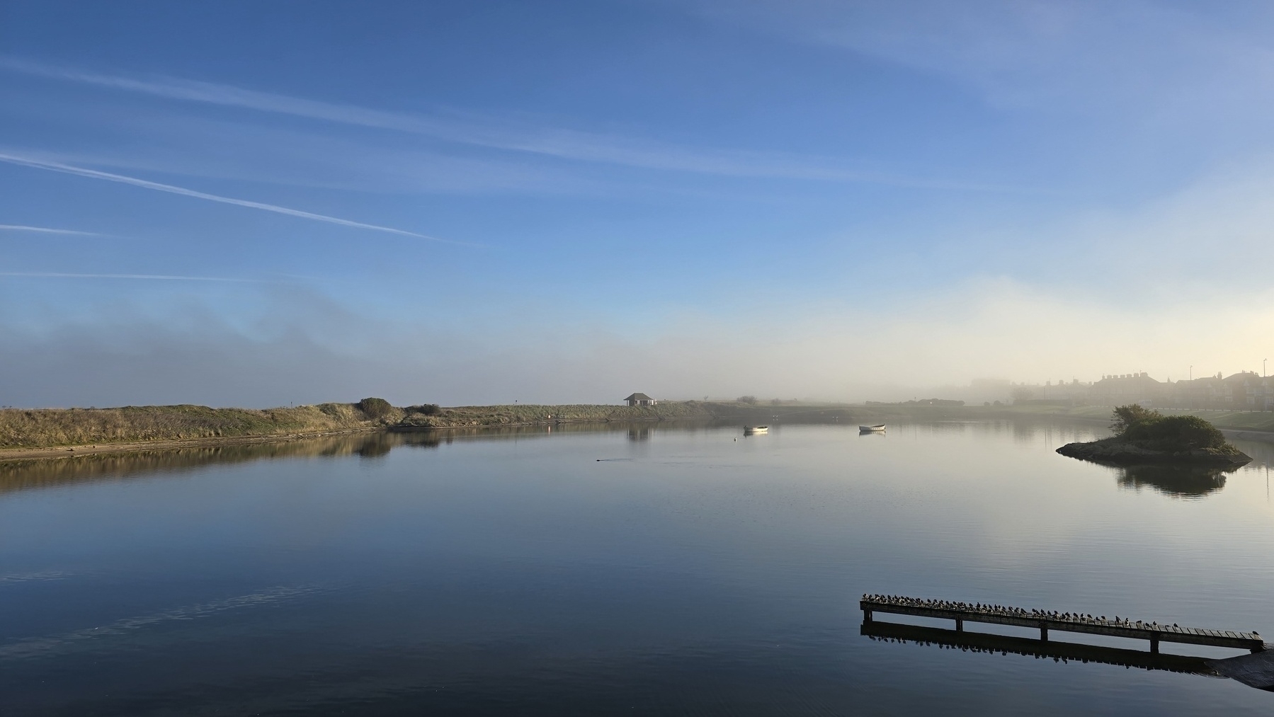 A misty boating lake, blue skies, a row of small birds line up on a jetty