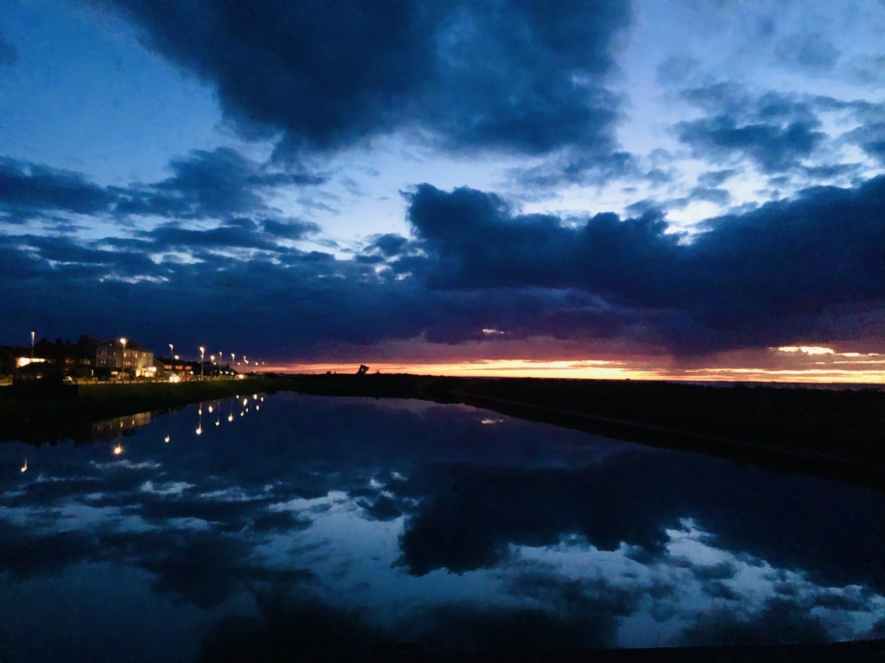 Sky and lake at sunset in a blue hue with a line of yellow on the horizon, the clouds reflect in the water along with a line of streetlights to the left of the frame