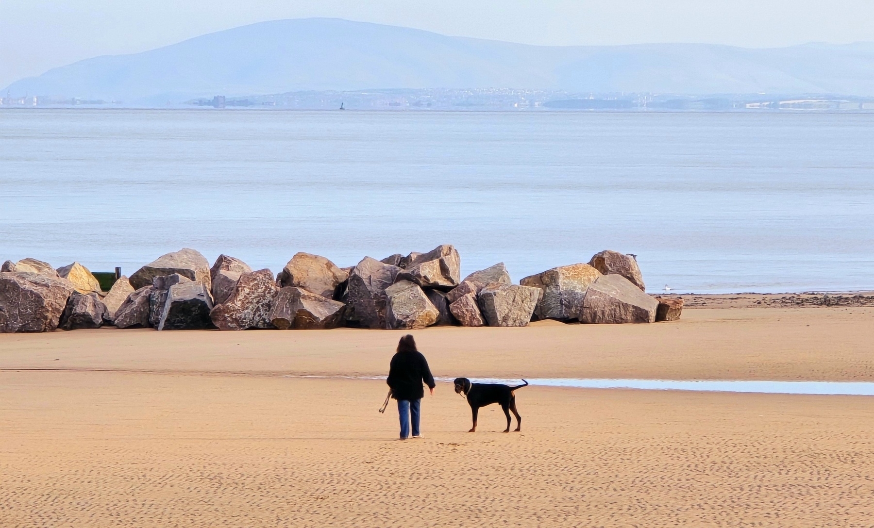 Beach scene with a lady walking her dog,  rocks in front of her,  mountains in the distance. 