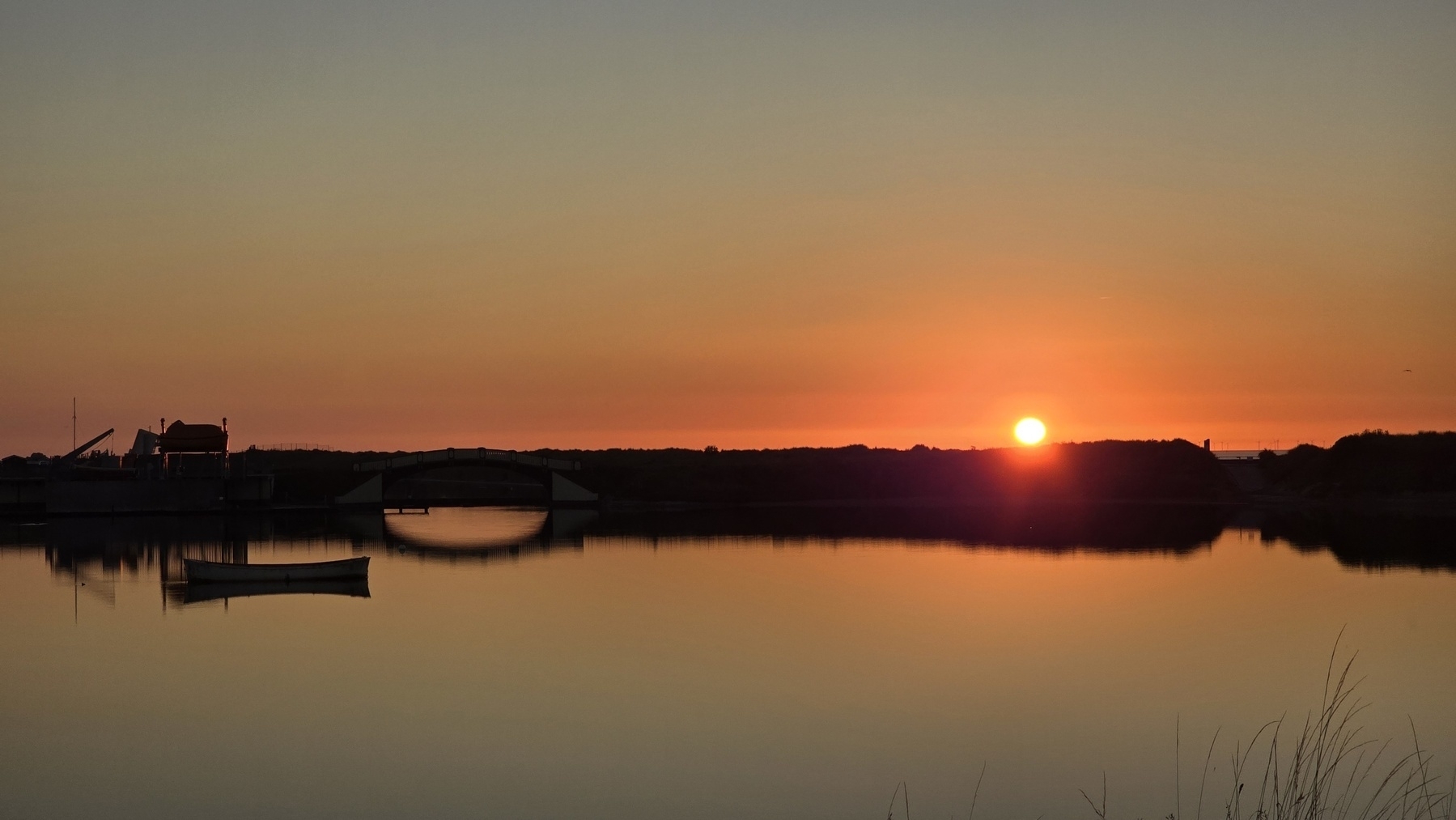 Sunset over a calm boating lake with orange sun resting on horizon, a bridge reflects off the water