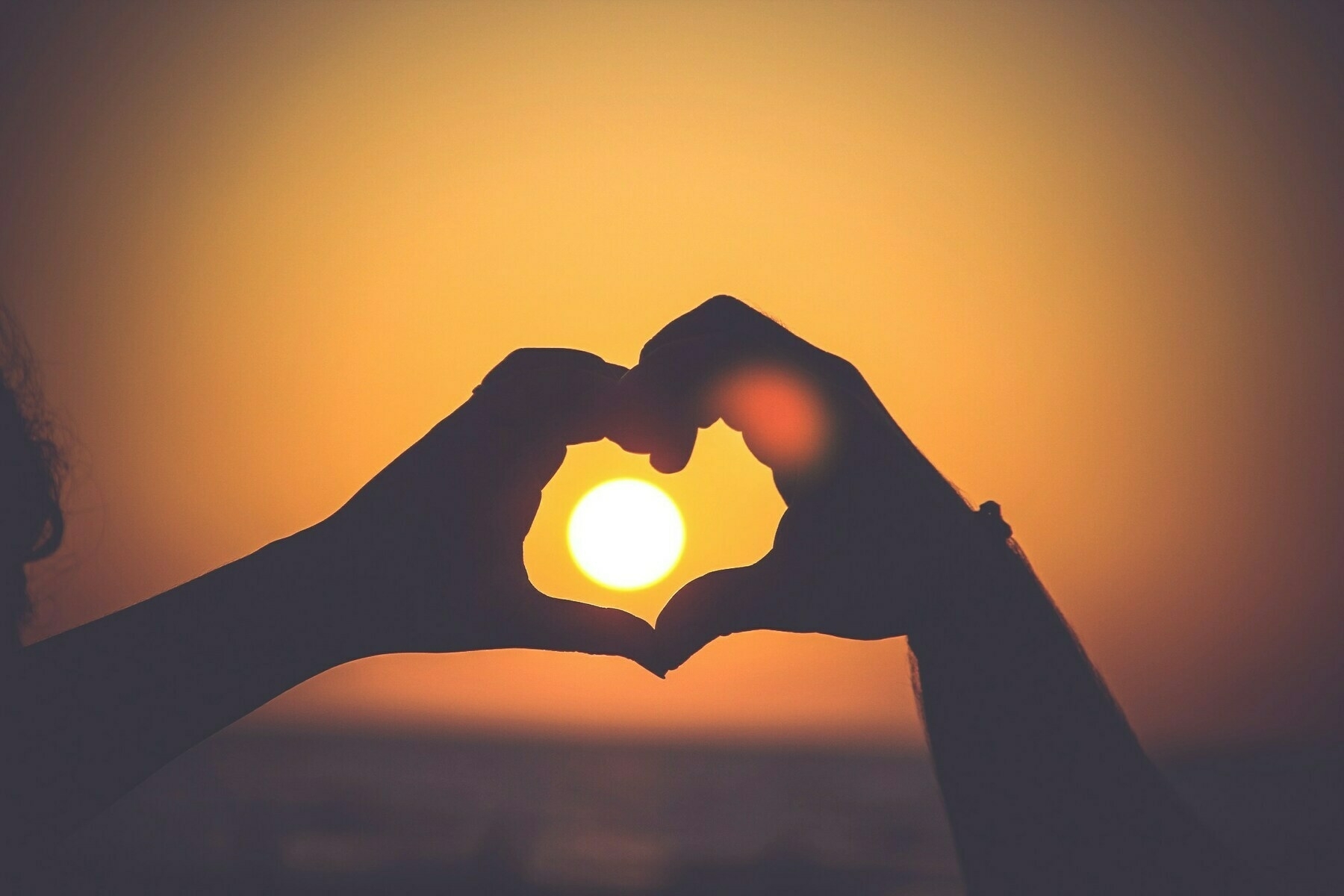 hands making a heart shape with a low, yellow sun framed in the background