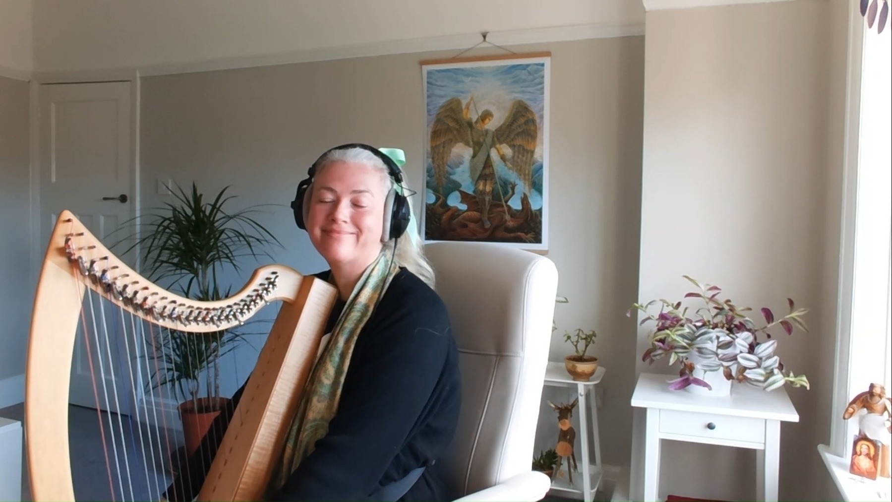 Holly holding her harp in her new studio filled with plants and a painting of Saint Michael on the wall behind her