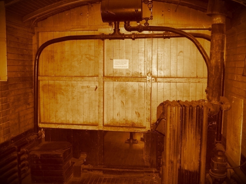 Sepia tone. Machinery and pipes. A wall of planks with an opening beneath. Brick walls on either side.