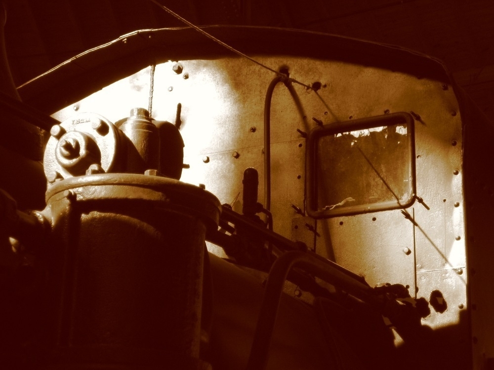 Sepia tone. Rivets and machinery at the front of a locomotive. A single window stares outward like an eye.