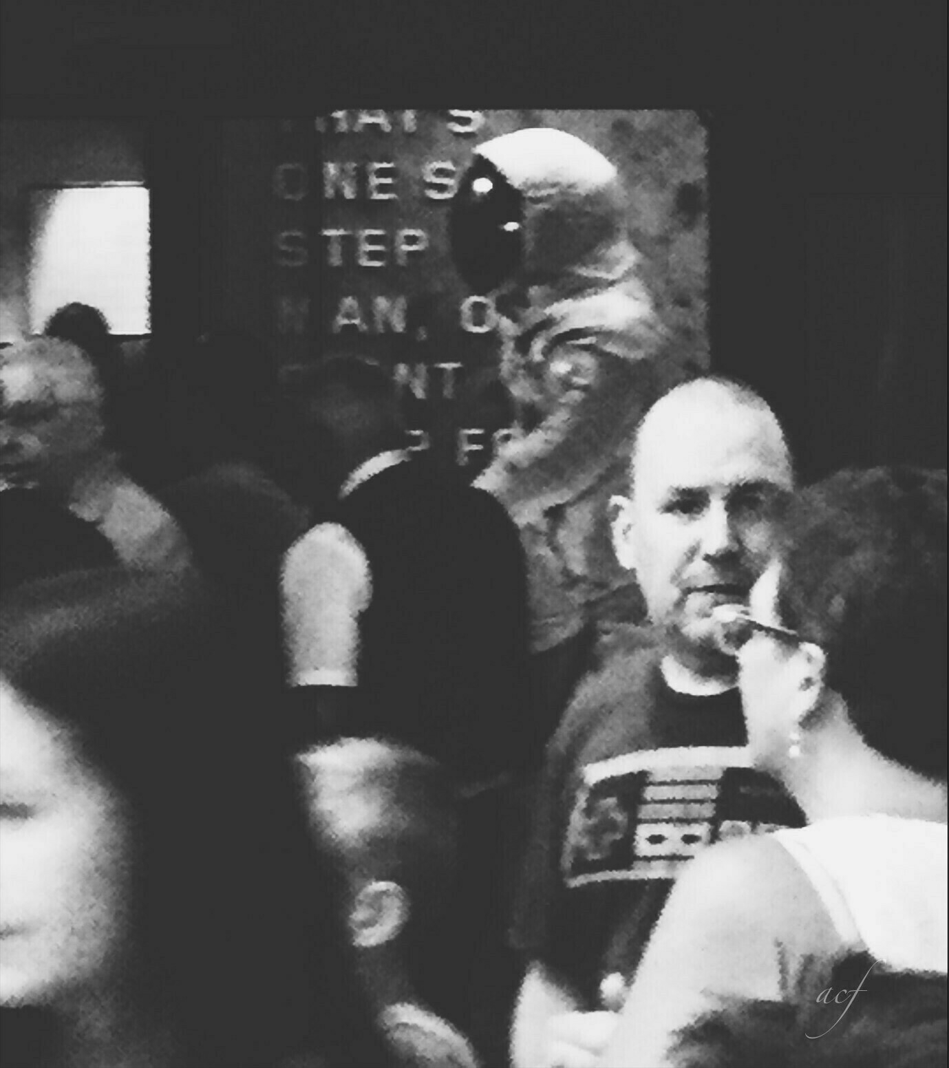 Black and white. A blurred crowd of people in foreground. Top of Armstrong spacesuit in background on display, facing left. The words 