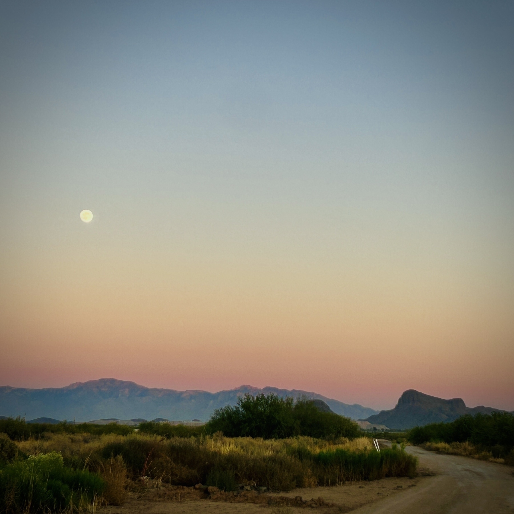 A dirt road leads into a desert landscape with a full moon in the sky and mountains in the background with a pink and blue sky.