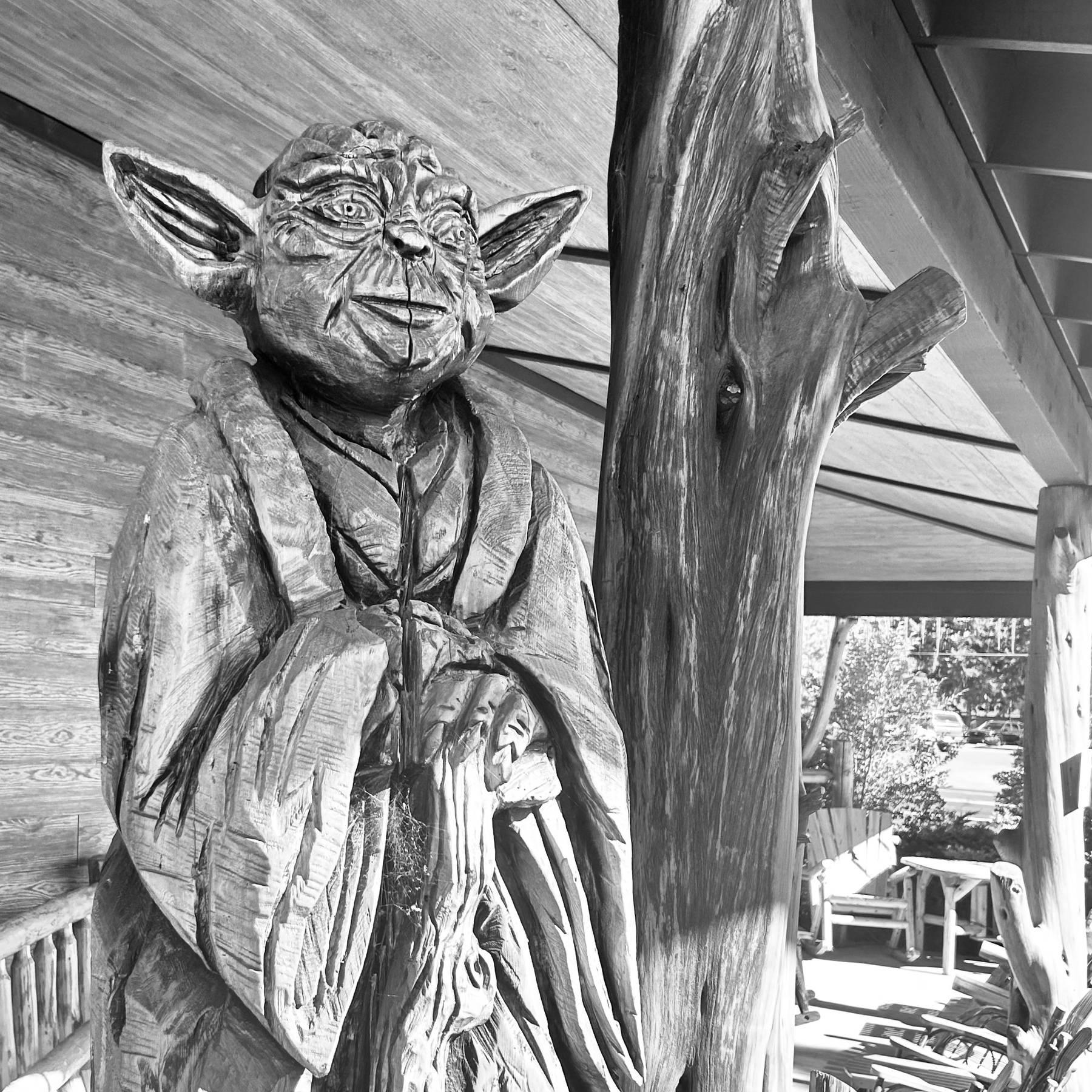 A wooden sculpture of Yoda stands next to a tree trunk and on a porch.