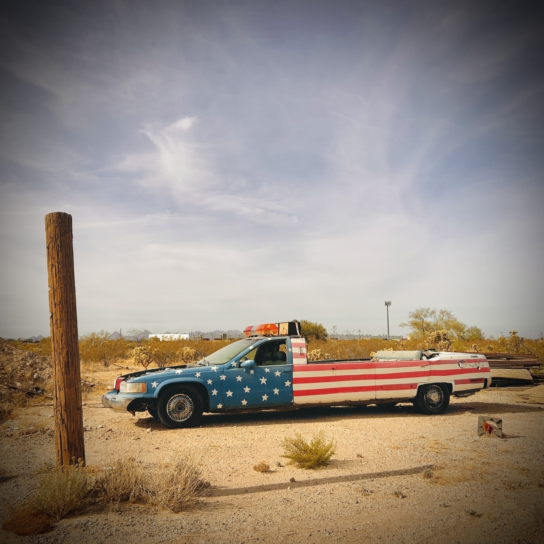 A former limousine truck painted with the American flag design is parked and decaying in a desert landscape. A cutoff telephone poll stands nearby. 