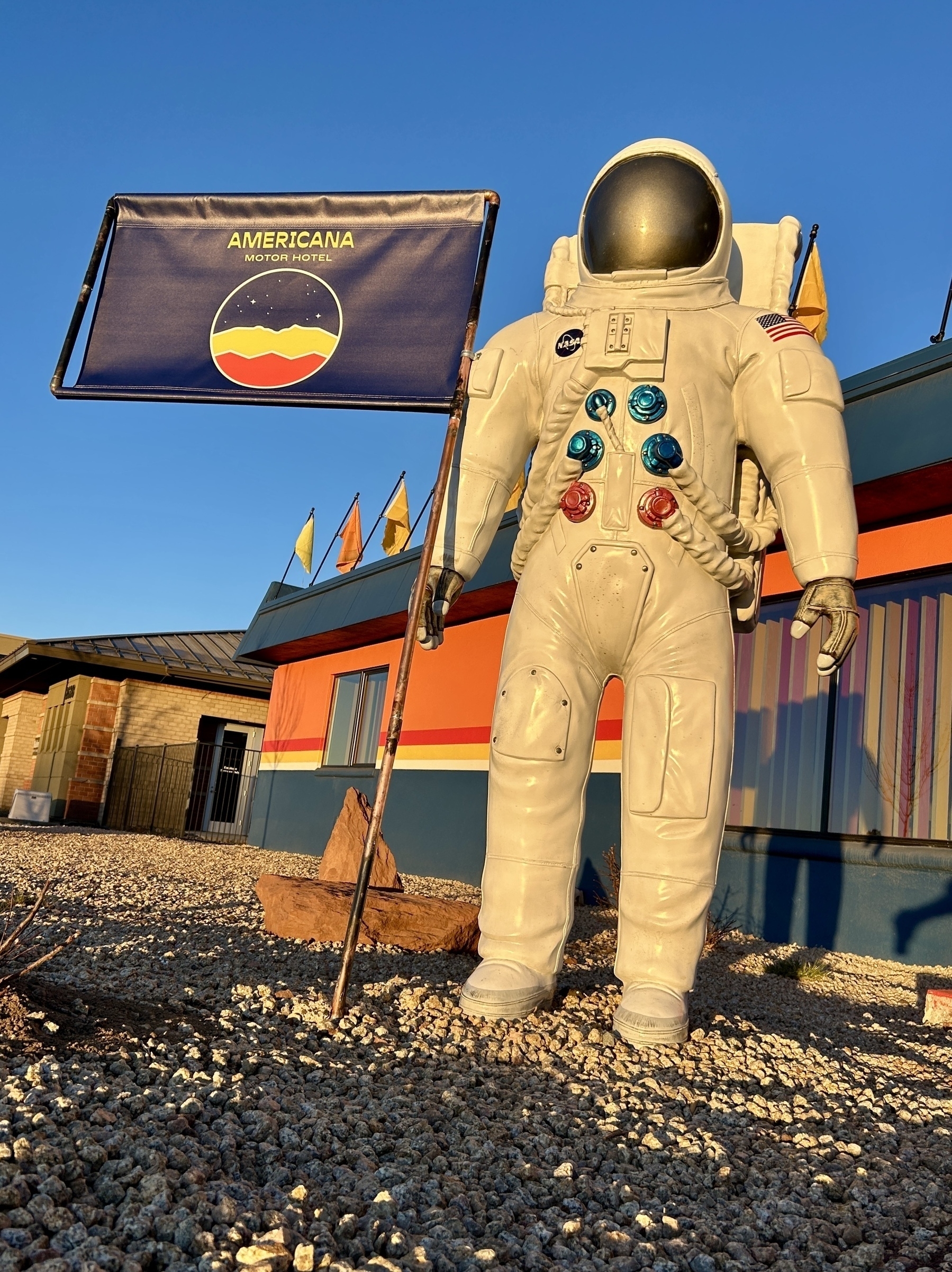 A statue of an astronaut holding a flag stands outside a colorful building with a sign reading "Americana Motor Hotel."