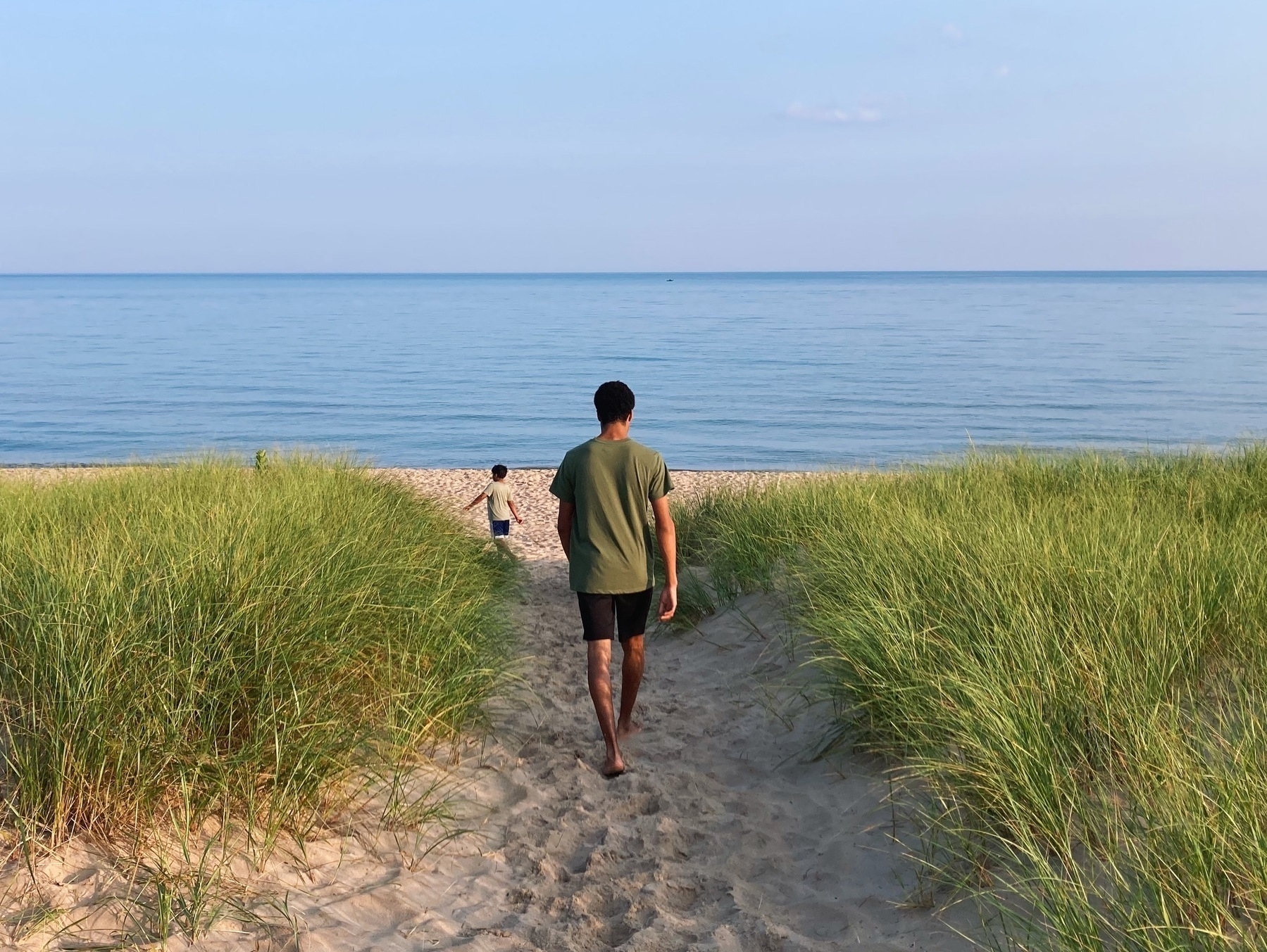 Two boys walking through grass on a sandy trail to the beach.