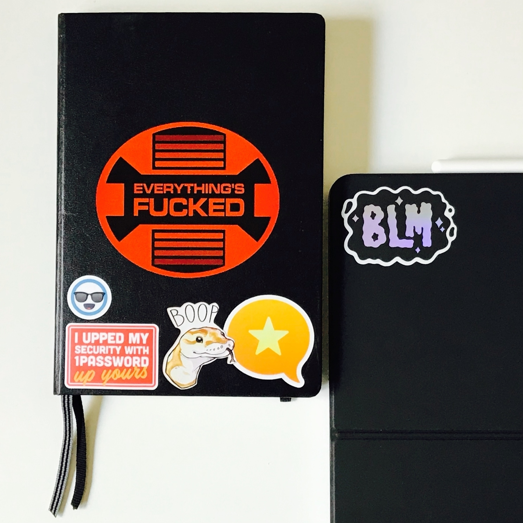 Bound black paper notebook with a few stickers on it. Next to it on the right, the top-left corner of an iPad Pro in a Magic Keyboard cover, a singular shiny BLM (Black Lives Matter) sticker on it.