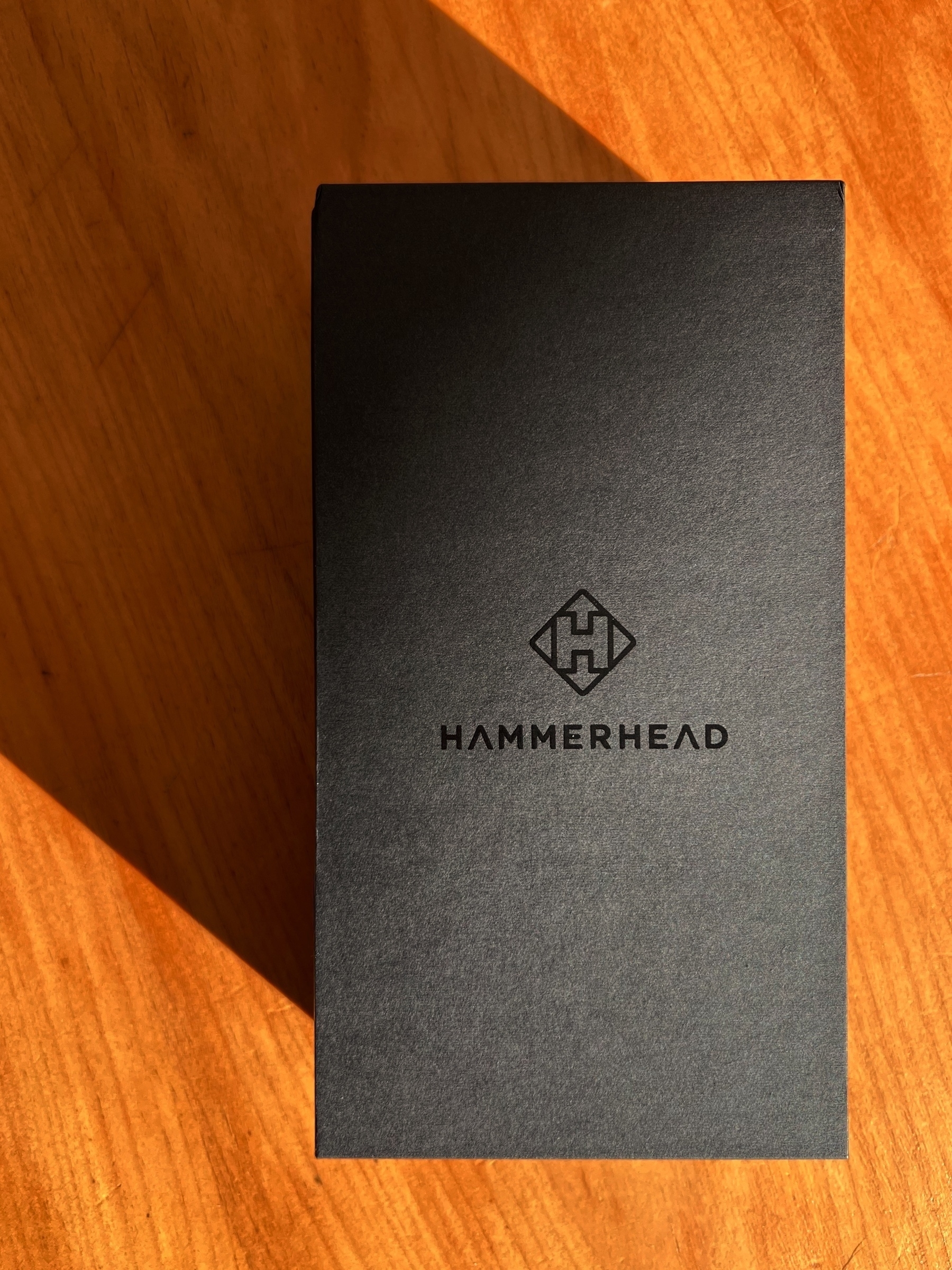 Rectangular paper packaging in matte black with a glossy ”Hammerhead“ brand name and logo in the middle sitting on top of a wooden table, sunlight harshly illuminating everthing from one side, casting a dark shadow to the left of the packaging.