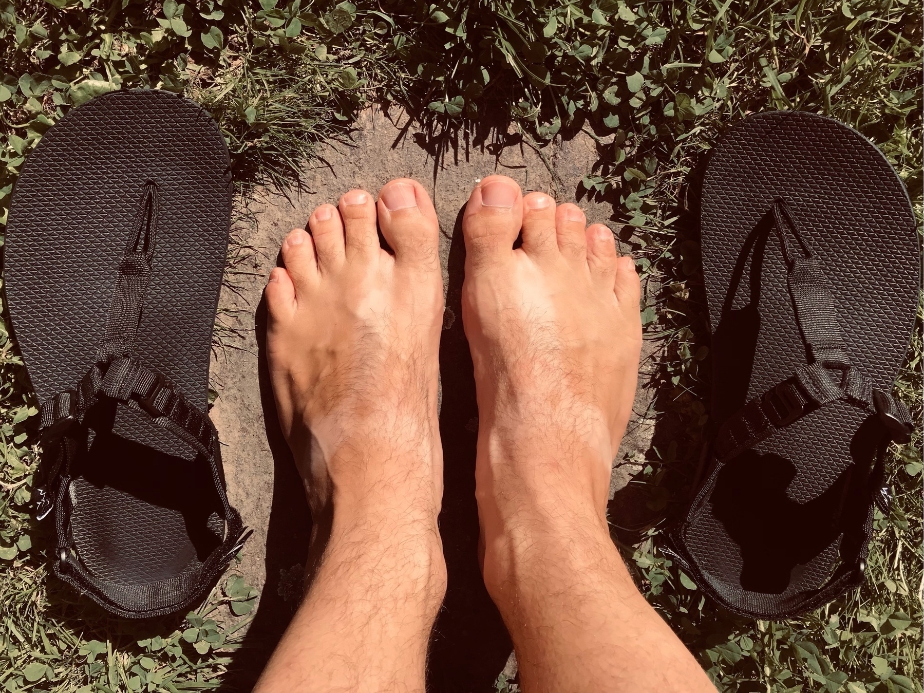 Top view of lightly tanned bare feet with tan lines in the shape of sandal straps. The black sandals that caused those tan lines laying to the left and right of the feet.