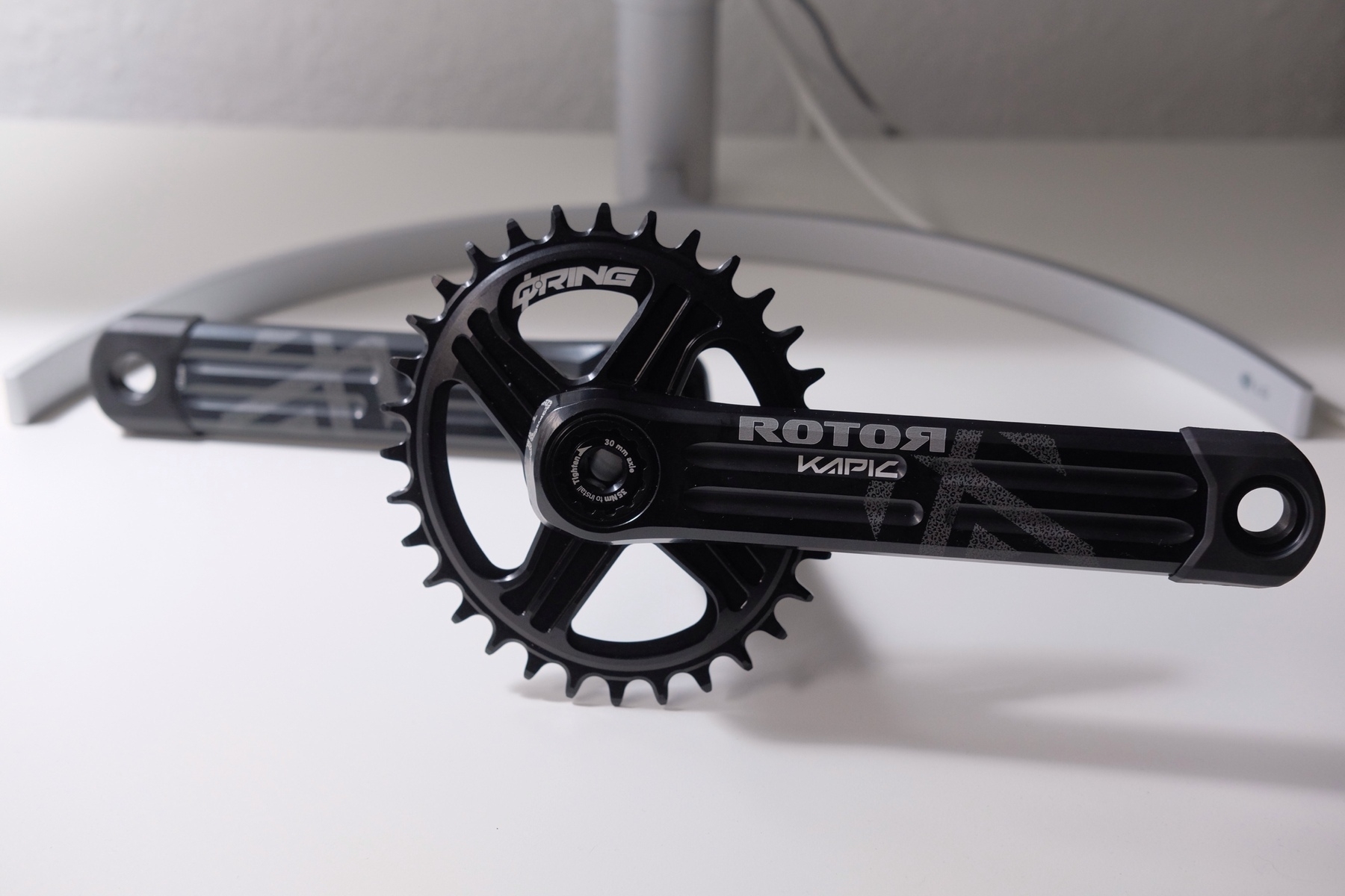 Aluminium single chainring crankset with an oval chainring on a white table in front of the foot of a monitor.