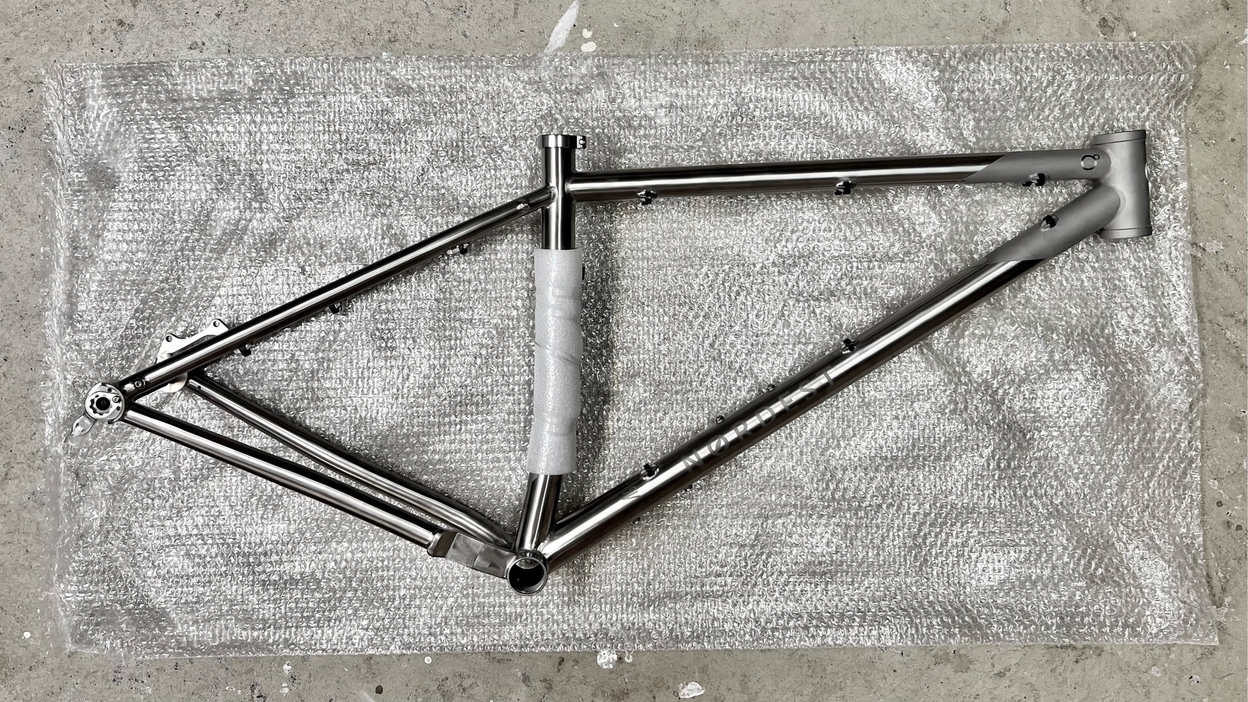 Brushed titanium mountain bike frame laying drive side up on a layer of bubble wrap on the ground. The seat tube is wrapped in a bit of cushioning plastic to hold the rear axle, which is presently out of view.